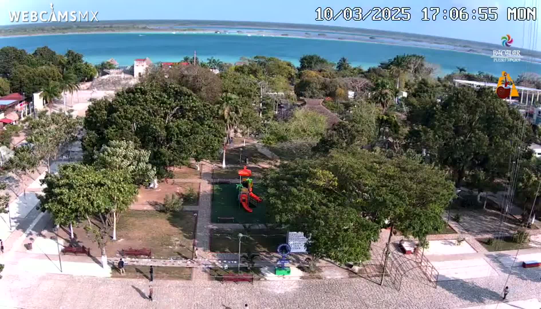 An aerial view shows a park with a playground next to a town overlooking a calm, turquoise body of water under a clear, sunny sky.