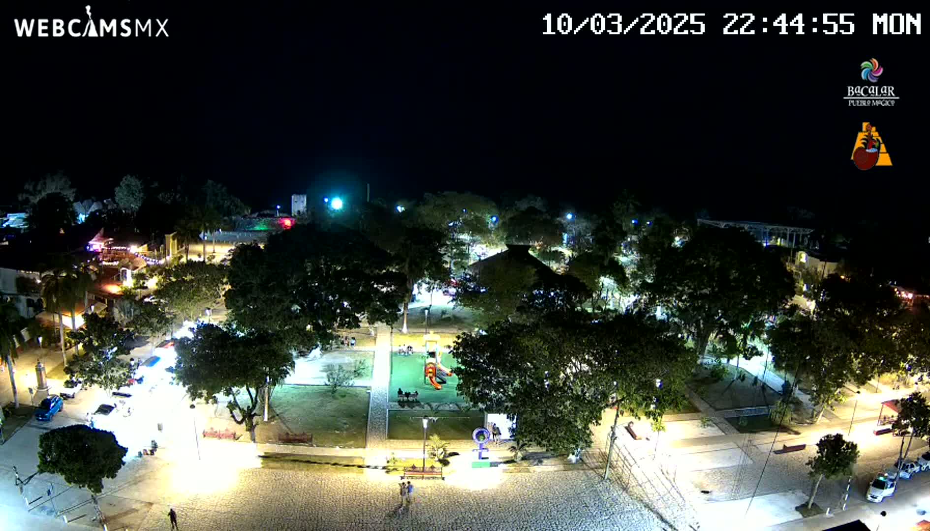 A nighttime aerial view of a town square with a playground, surrounded by trees and buildings, illuminated by streetlights under a clear sky.