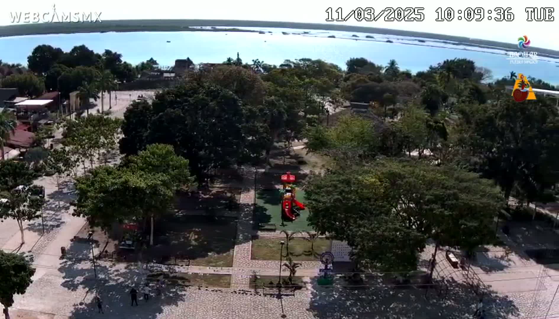 An aerial view shows a park with a playground near a body of water on a sunny day.