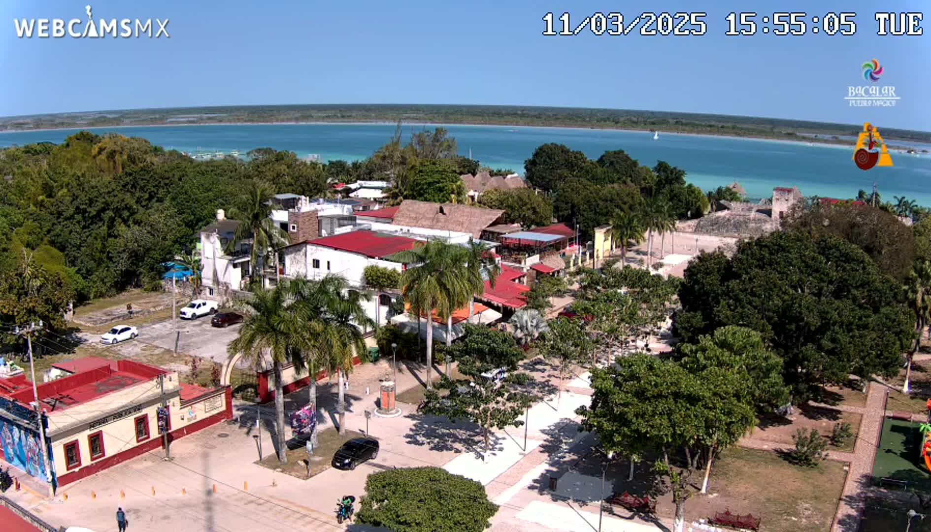 An aerial view of a town square with red-roofed buildings, palm trees, and a turquoise body of water in the background on a sunny day.