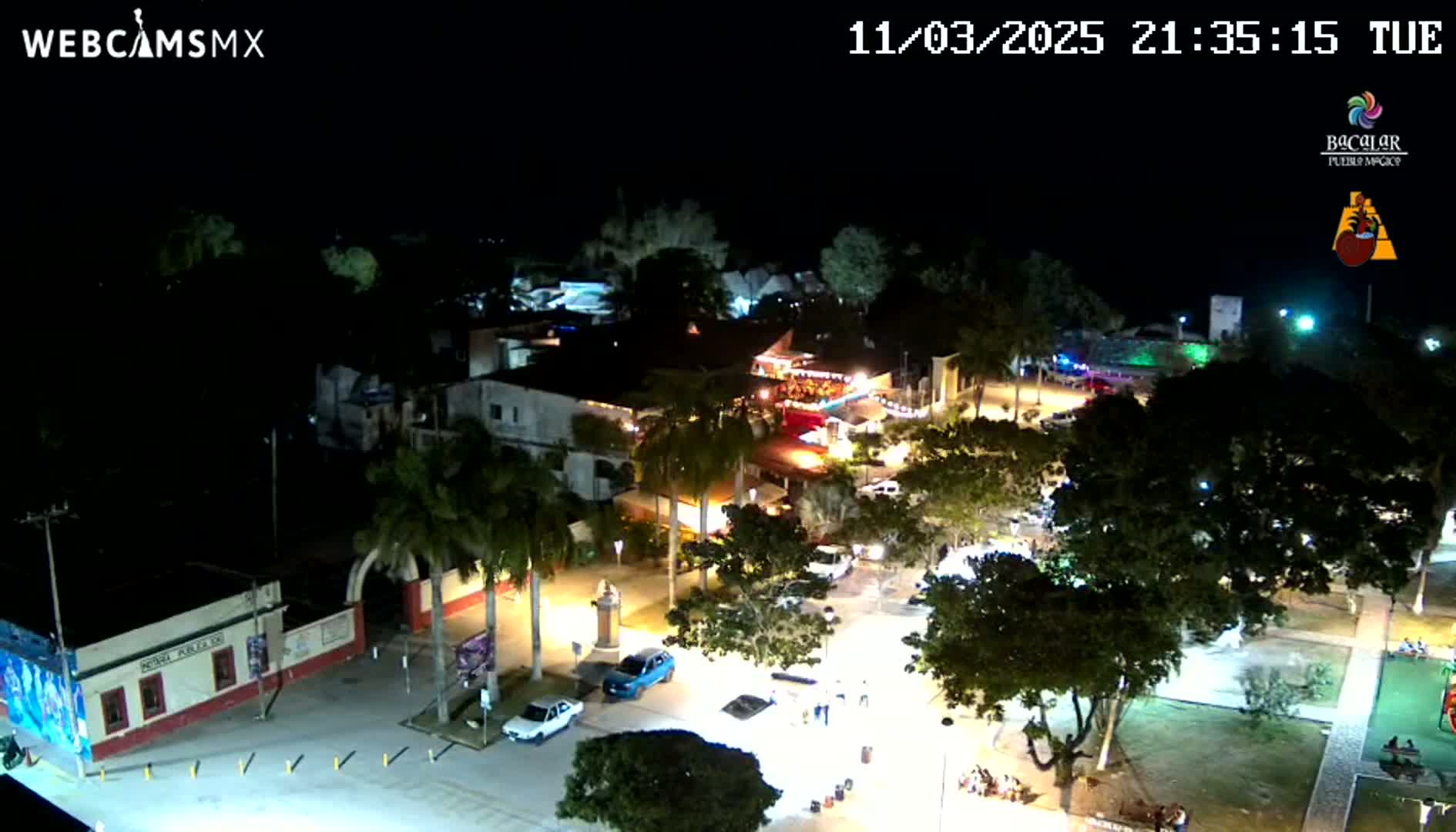 A nighttime, aerial view of a town square with buildings, trees, cars, and people illuminated by artificial light.