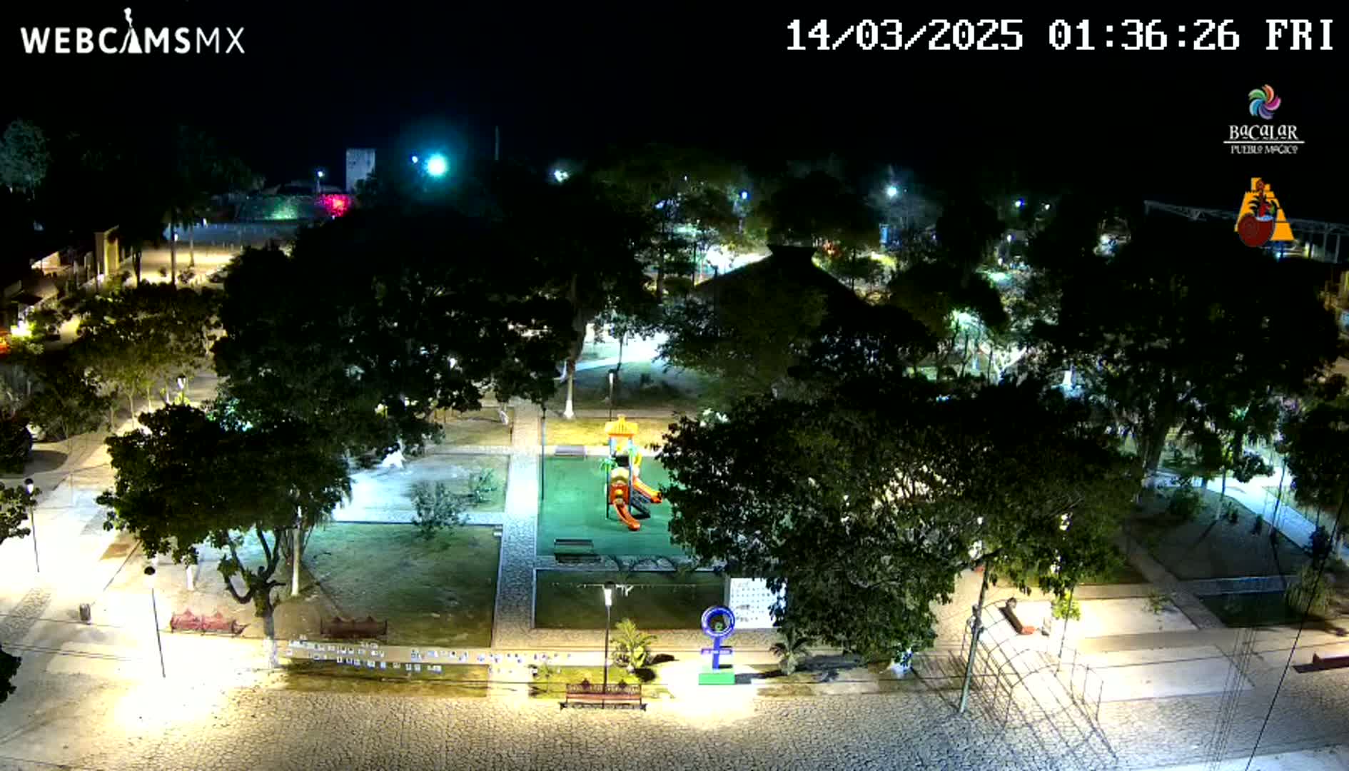 A nighttime aerial view of a park with a playground, surrounded by trees and paved walkways, illuminated by artificial lighting.