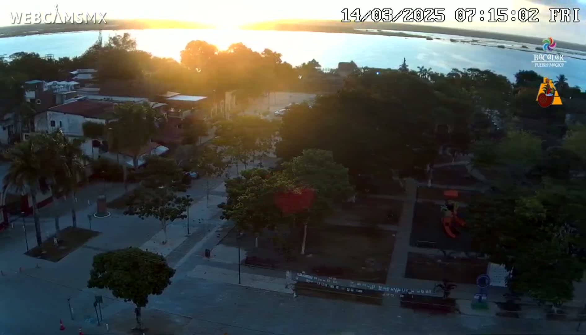 An aerial view of a town square at sunrise, showing buildings, trees, a playground, and a body of water in the background.