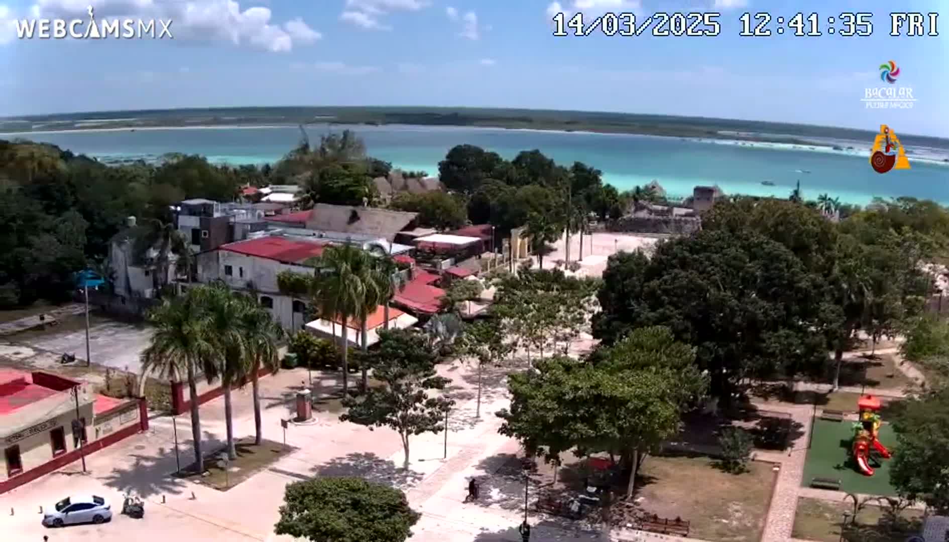 An aerial view shows a town square with buildings, palm trees, a playground, and a park, overlooking a calm, turquoise body of water under a partly cloudy sky.