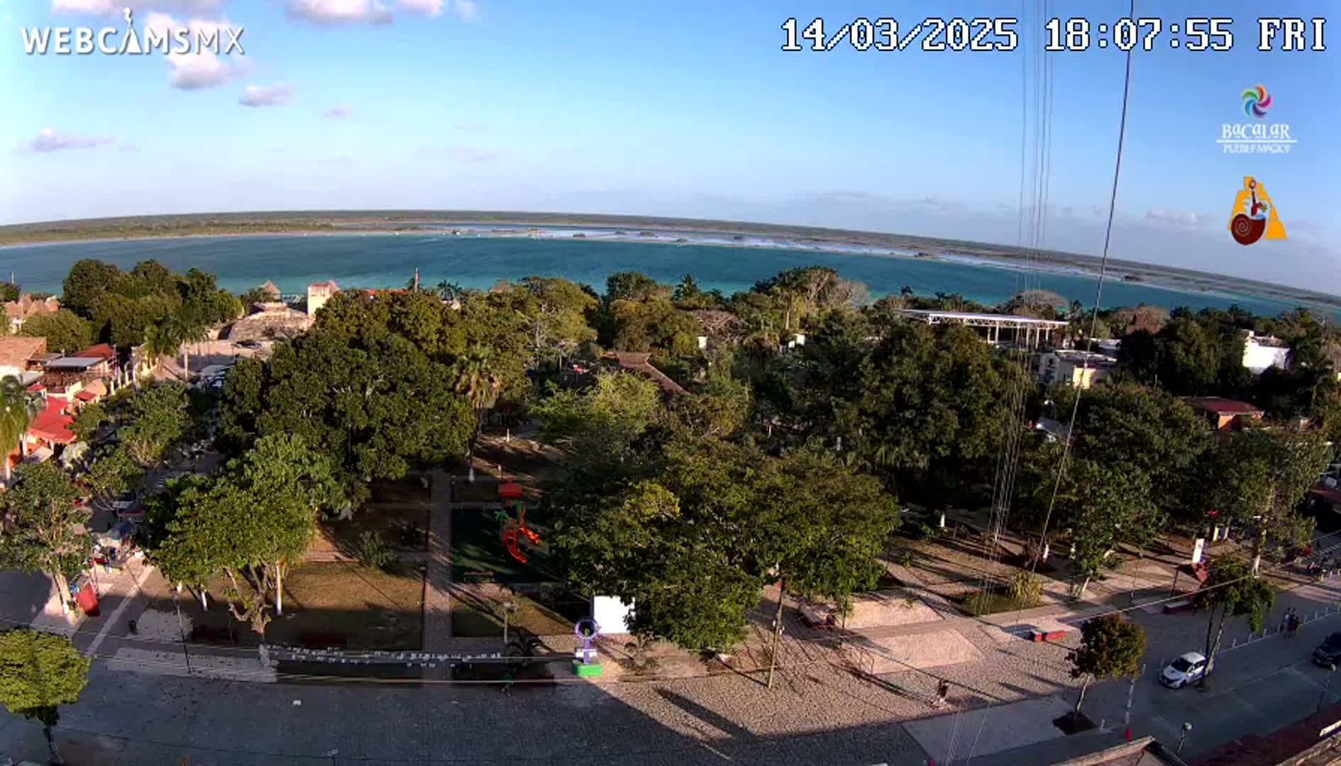 An aerial view of a town with a park and playground near a turquoise body of water under a mostly clear sky.