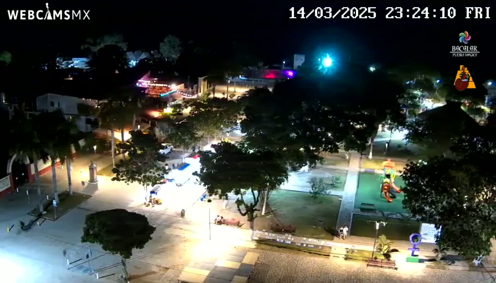 A nighttime aerial view of a town square with illuminated buildings, trees, and a playground, under a clear sky.