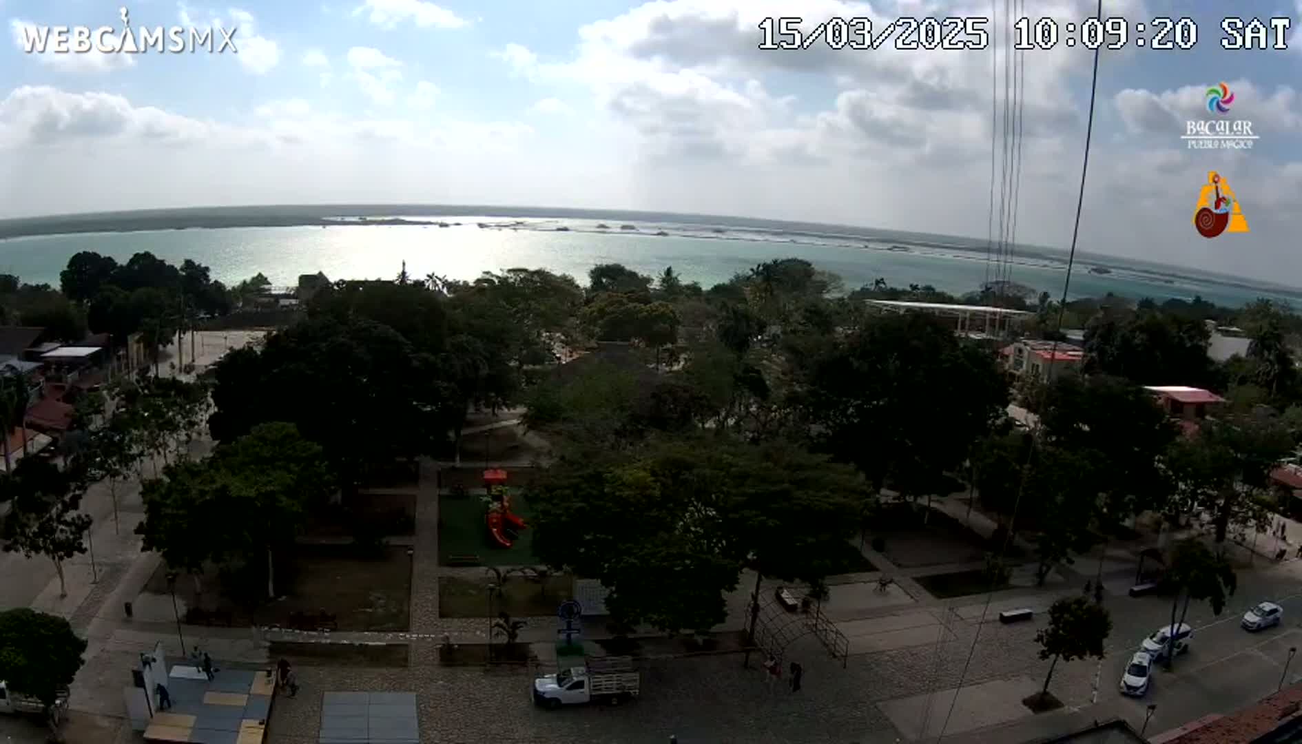 An elevated view shows a town square with a playground, trees, and parked cars, overlooking a calm body of water under a partly cloudy sky.