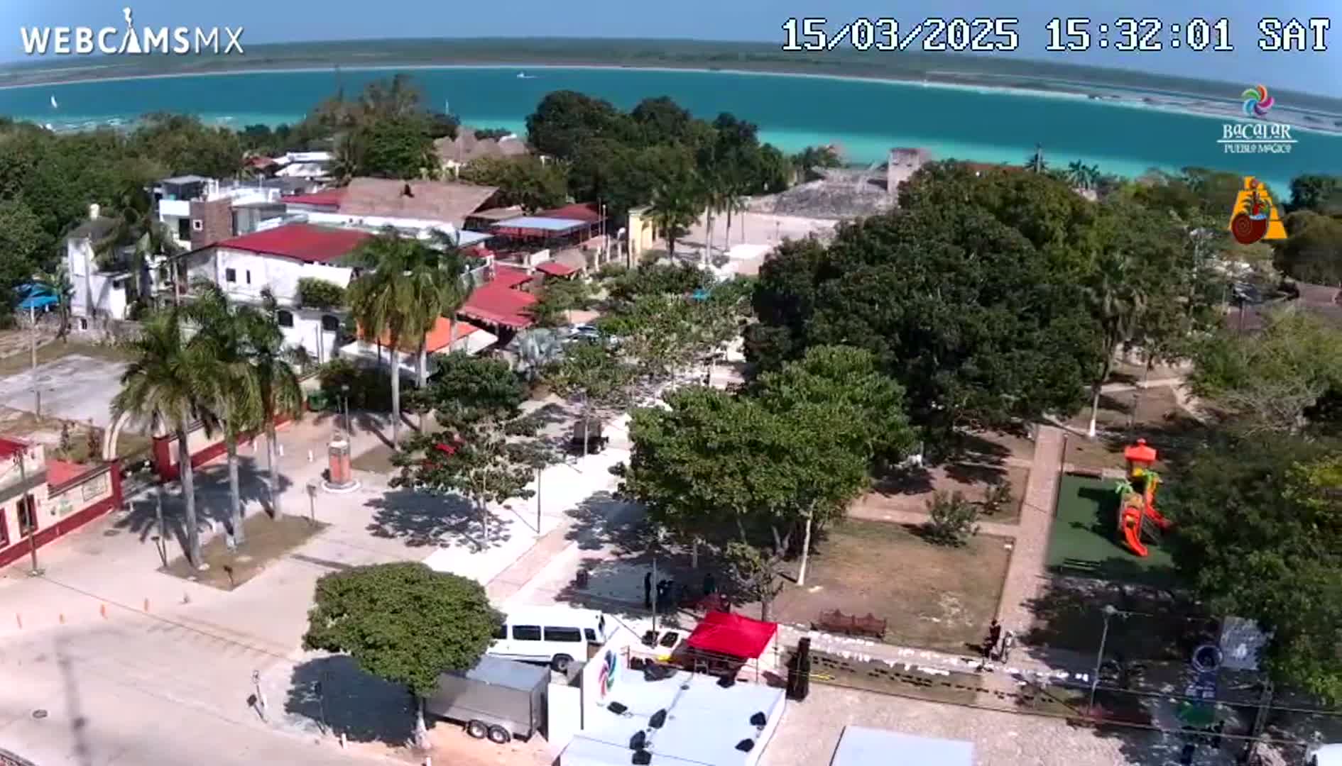 An aerial view shows a town's plaza with buildings, palm trees, a playground, and a turquoise body of water in the background under sunny skies.