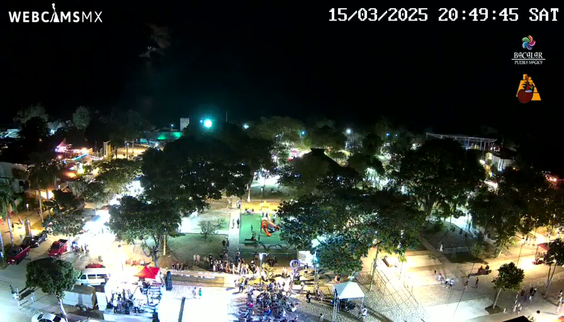 An illuminated town square at night, filled with people and trees, under a clear, dark sky.