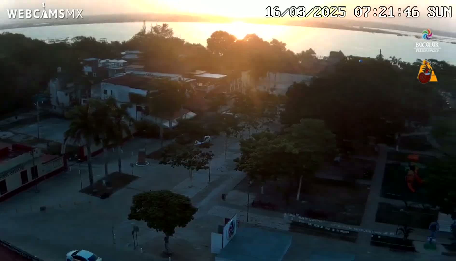An aerial view of a town square at sunrise, overlooking a calm body of water in the distance.