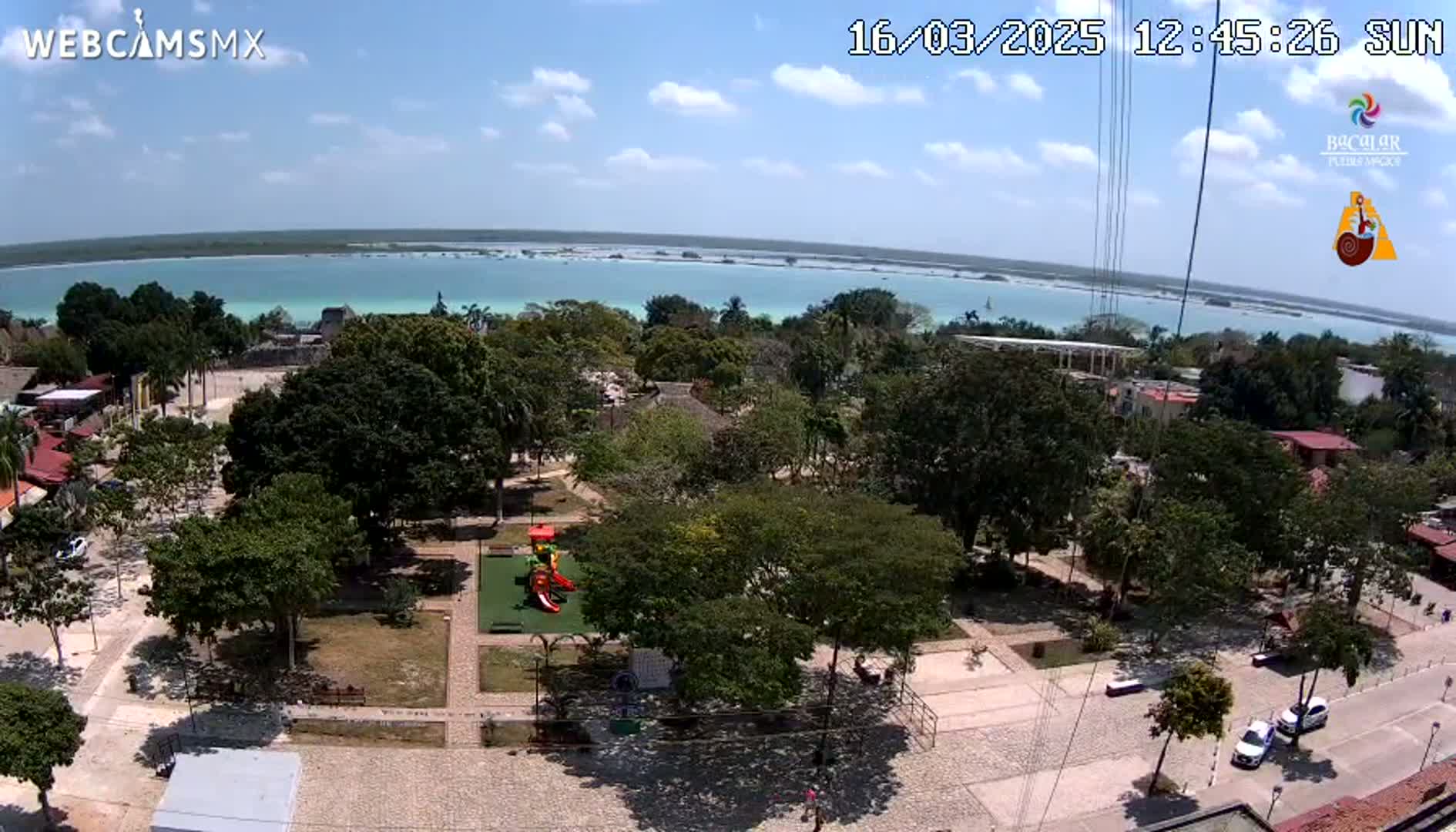 An aerial view of a town square with a playground, surrounded by lush green trees and buildings, overlooking a calm turquoise body of water under a sunny sky.