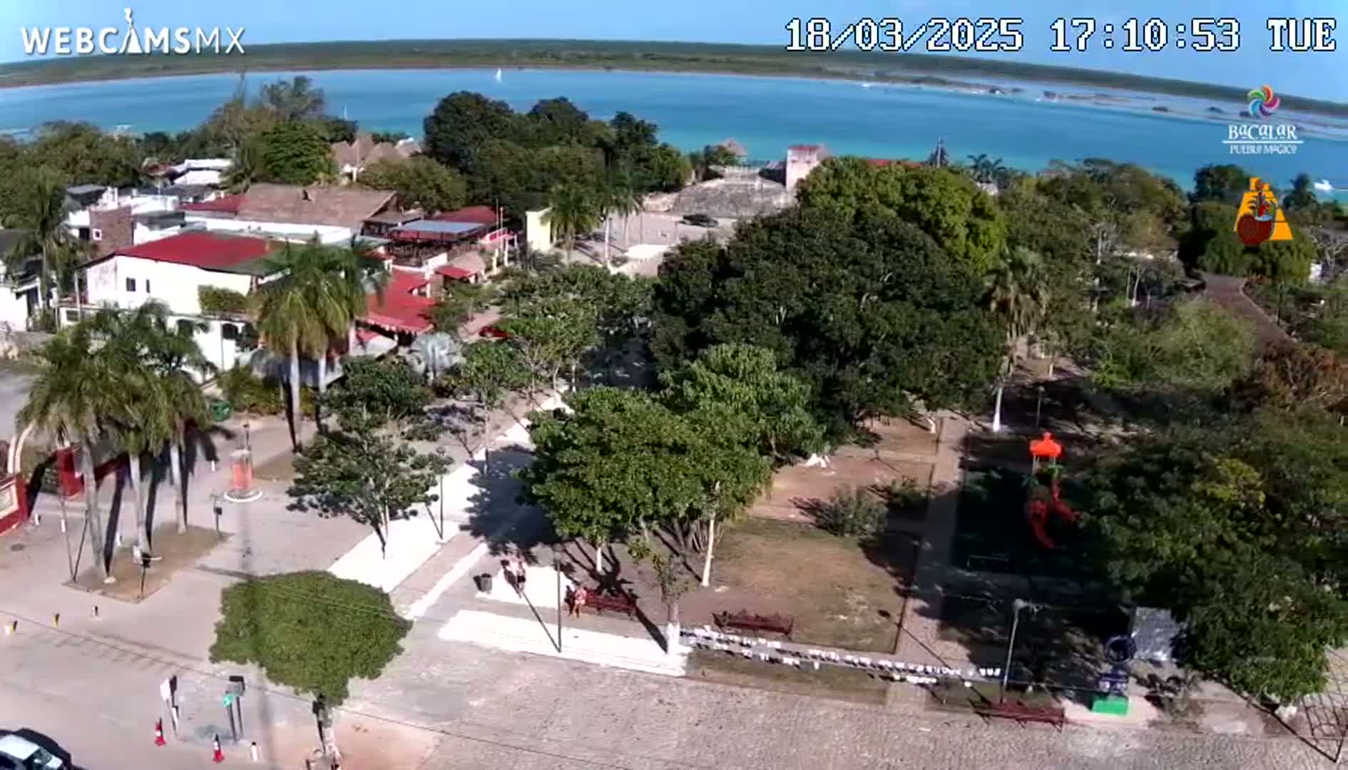 An aerial view shows a town square with palm trees, buildings, and a park next to a body of calm blue water under a sunny sky.