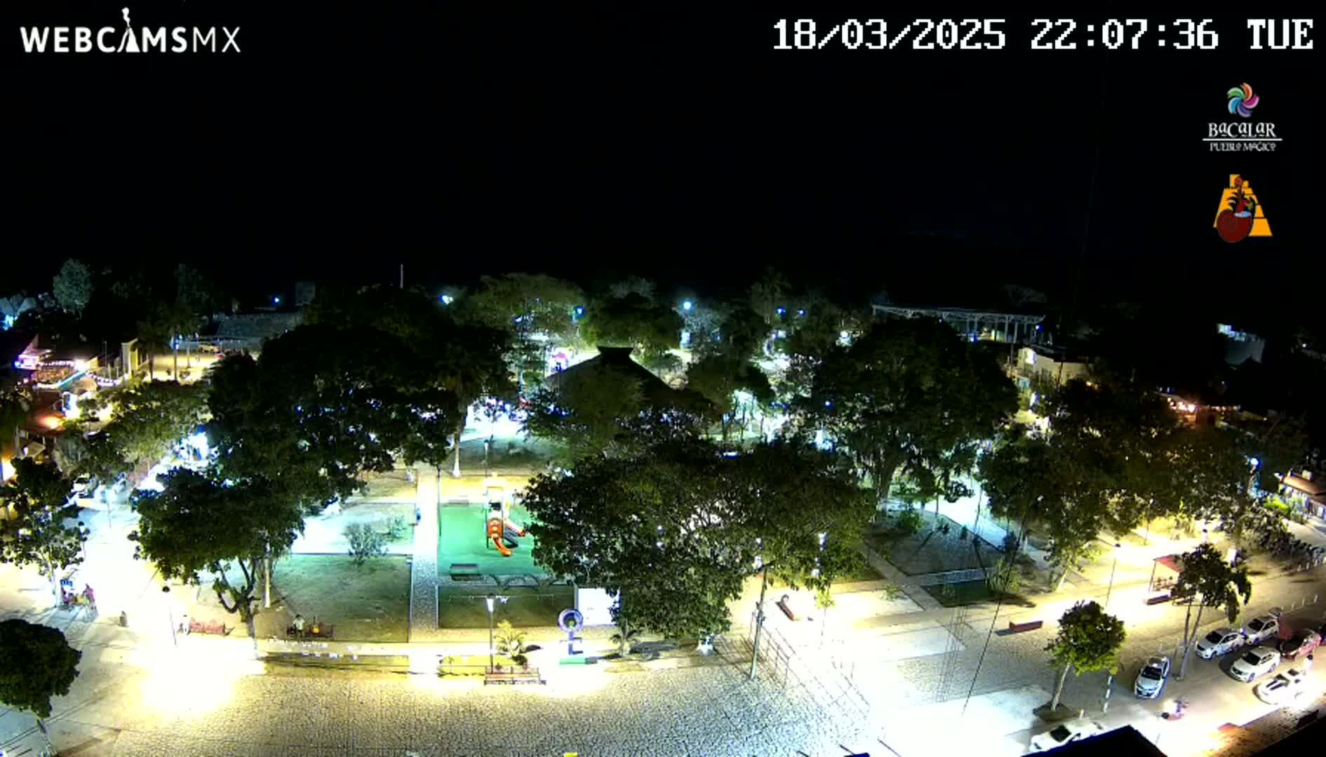 A nighttime aerial view of a town square with numerous trees, lights, and a few parked cars under a clear sky.