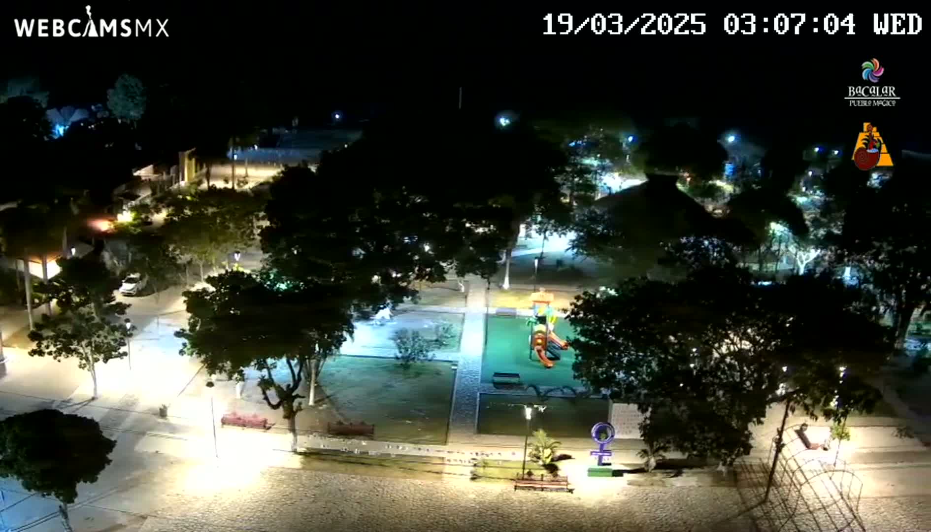 An illuminated nighttime view of a park with trees, pathways, and playground equipment.