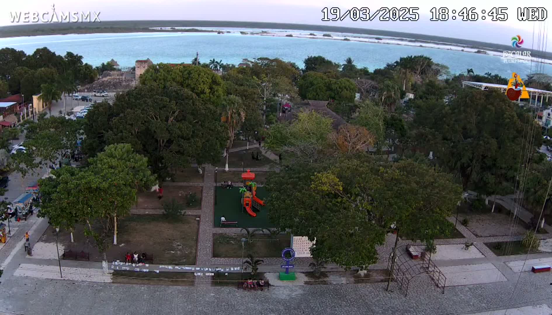 An aerial view of a town square with a playground, surrounded by lush green trees, overlooking a calm body of water under a twilight sky.