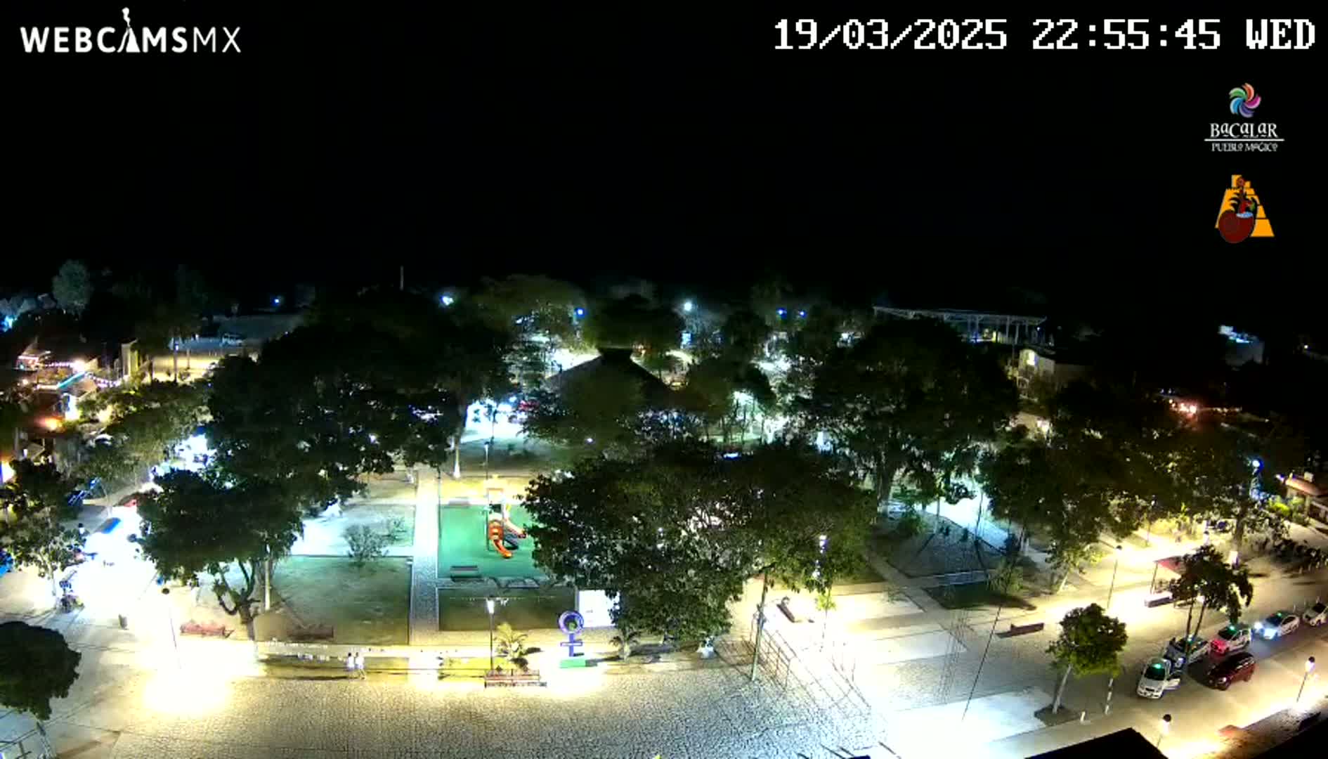 An illuminated town square at night, filled with trees and a playground, under a clear, dark sky.