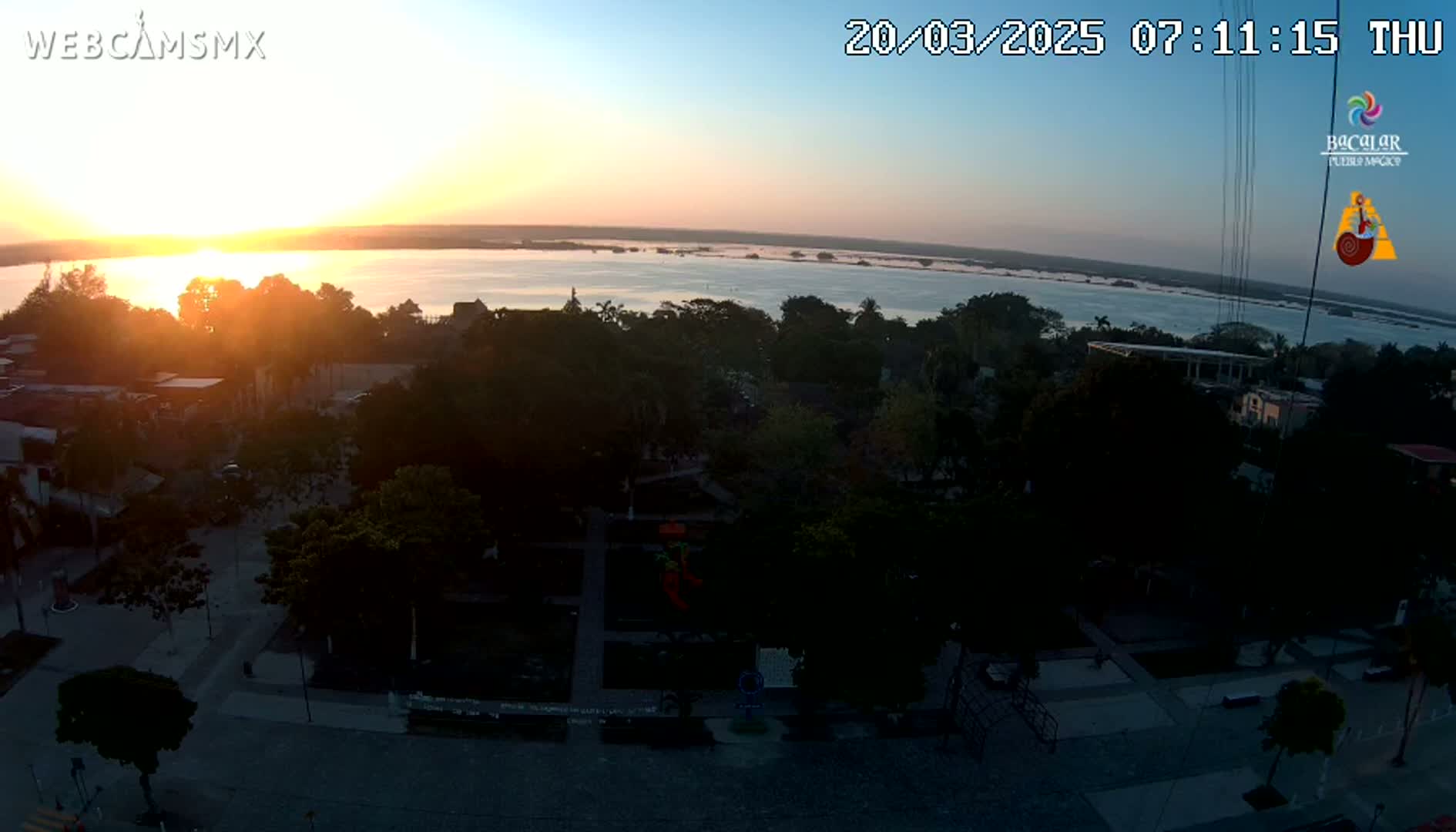 A sunrise over a calm body of water, seen from an elevated vantage point overlooking a town square with lush trees and buildings.