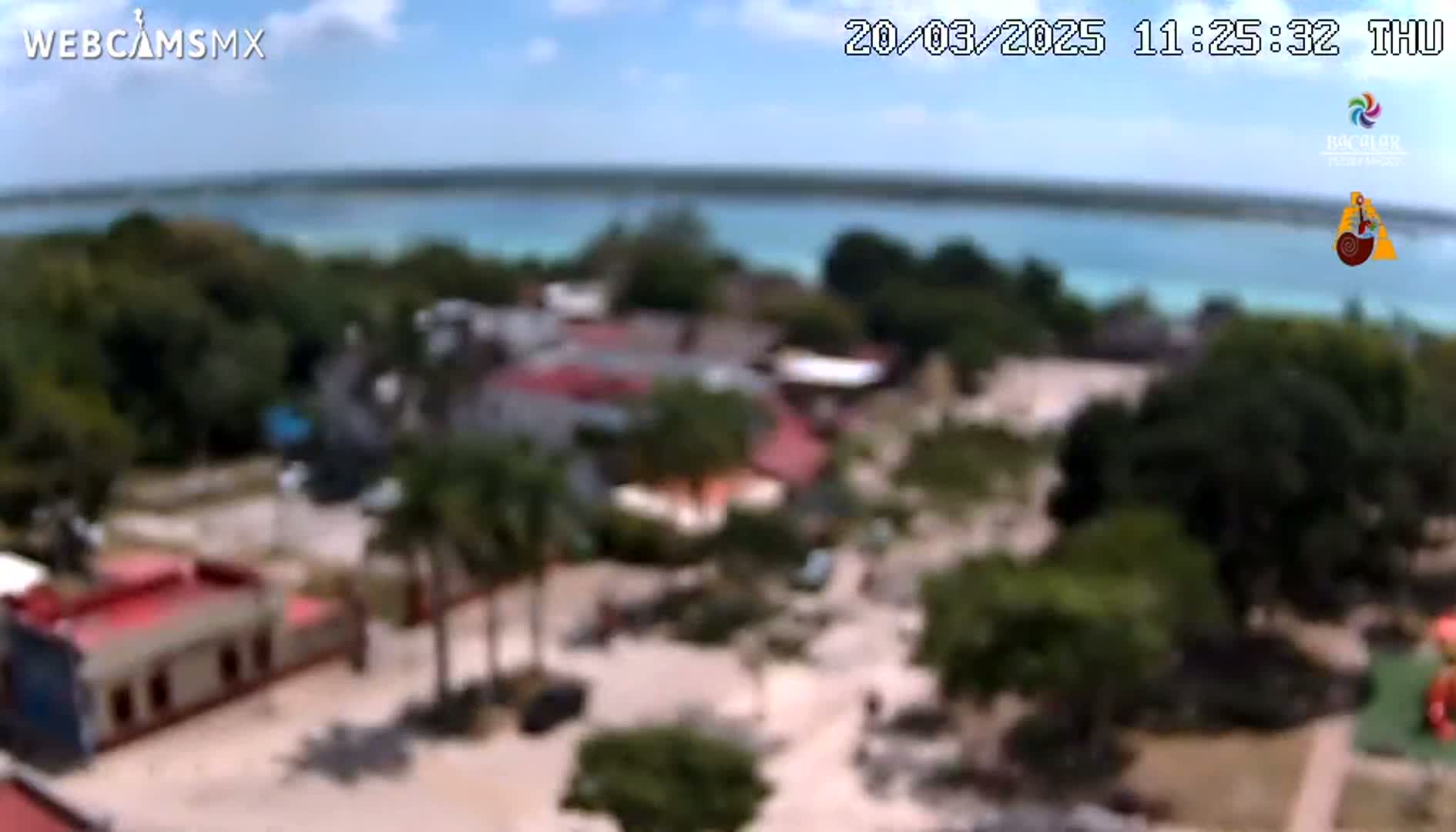 An elevated view of a coastal town with buildings and lush green vegetation under a partly cloudy sky, overlooking a calm body of turquoise water.