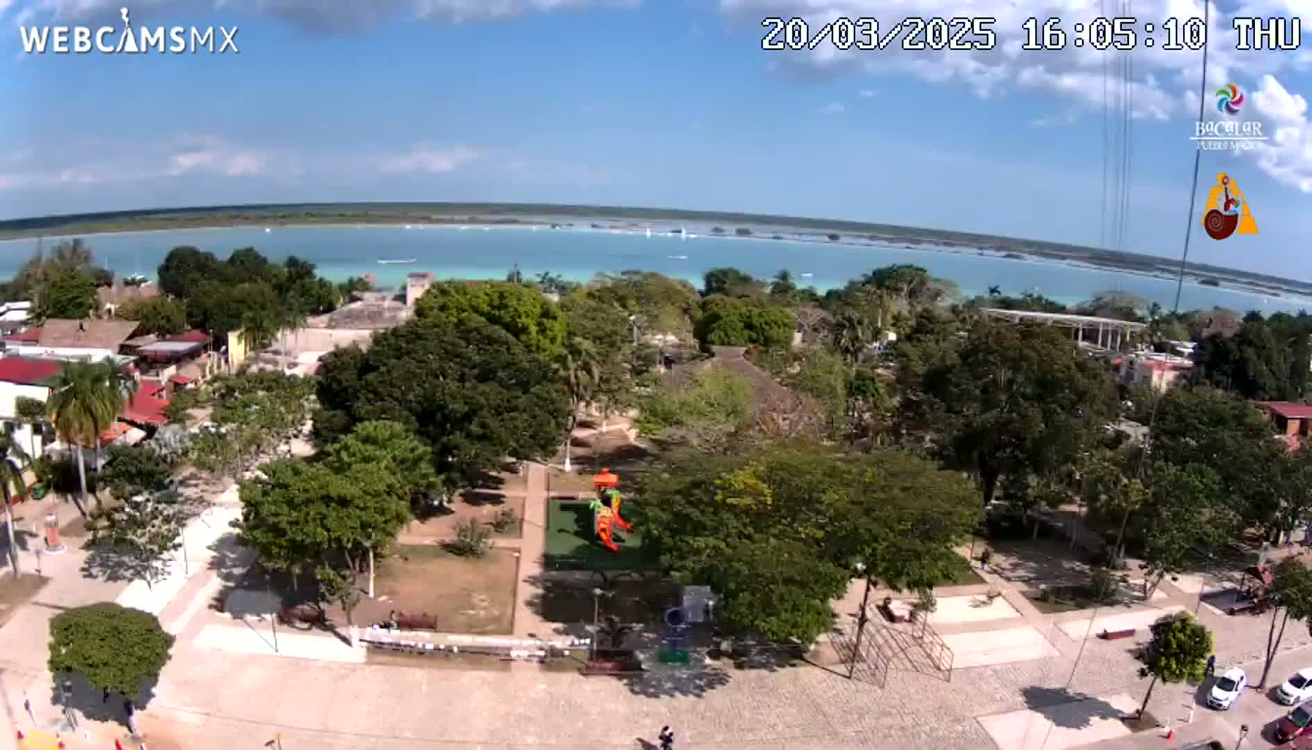 An aerial view of a town square with lush green trees and a playground, overlooking a calm, turquoise body of water under a partly cloudy sky.