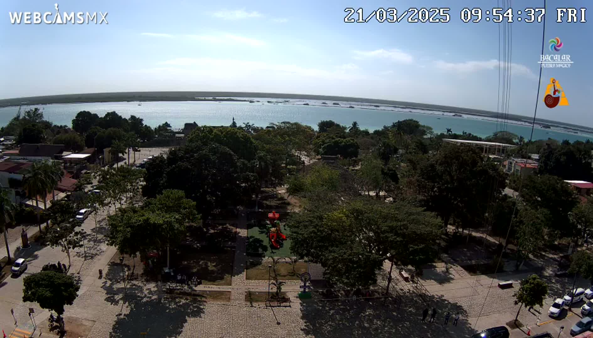 An aerial view shows a town square with a playground and surrounding trees, overlooking a calm, light-blue body of water under a sunny sky.