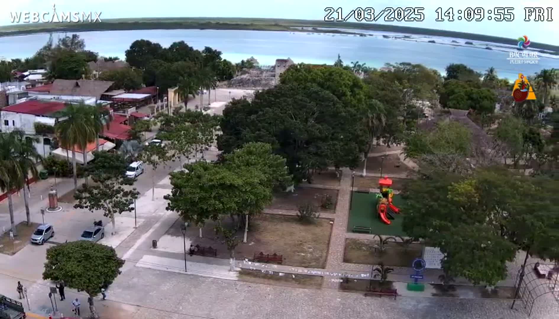 An aerial view shows a town square with a playground,  surrounding buildings, and a body of water in the background under a mostly clear sky.