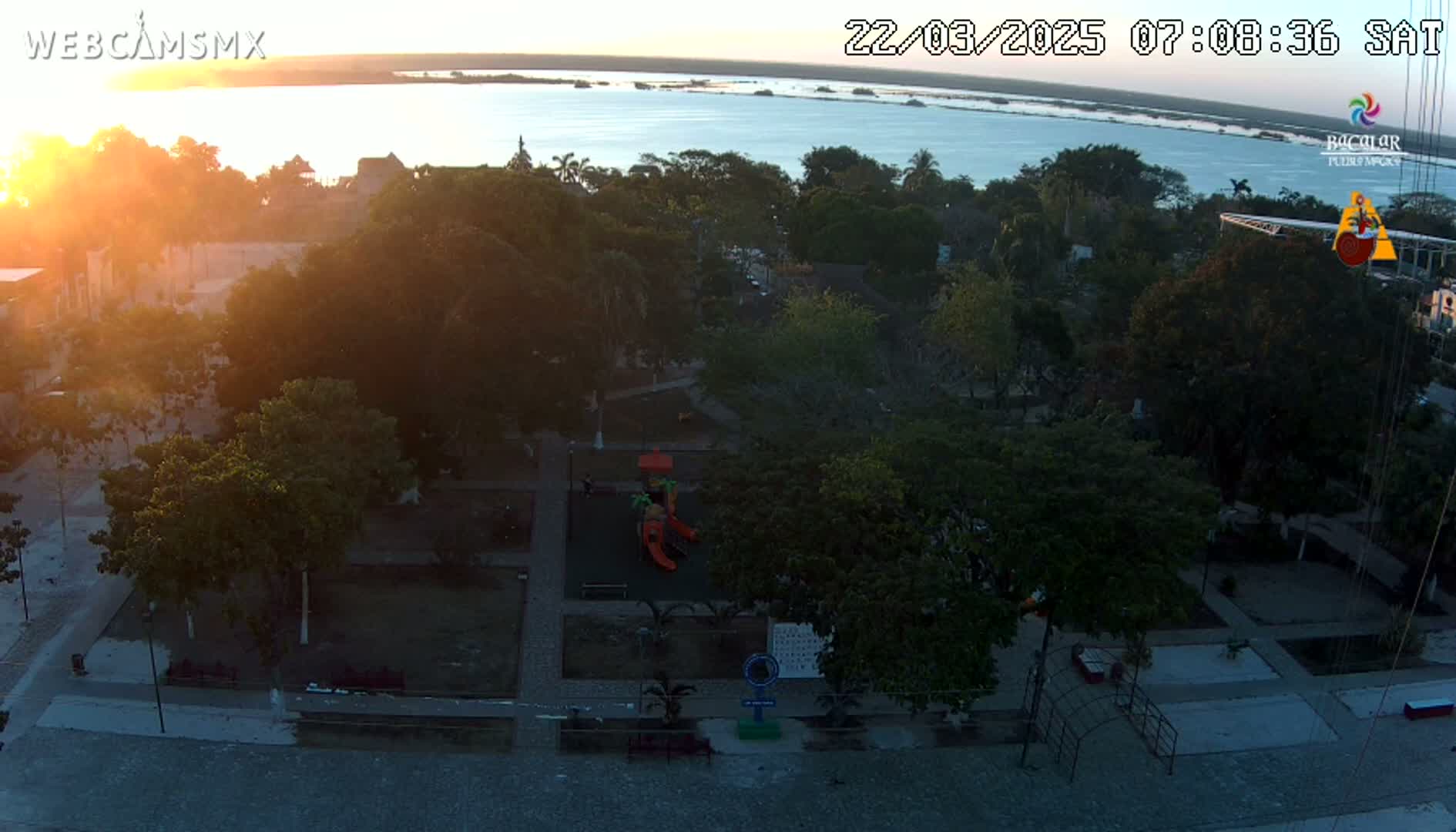A high-angle, sunset view of a town square with a playground, many trees, and a calm body of water in the background.