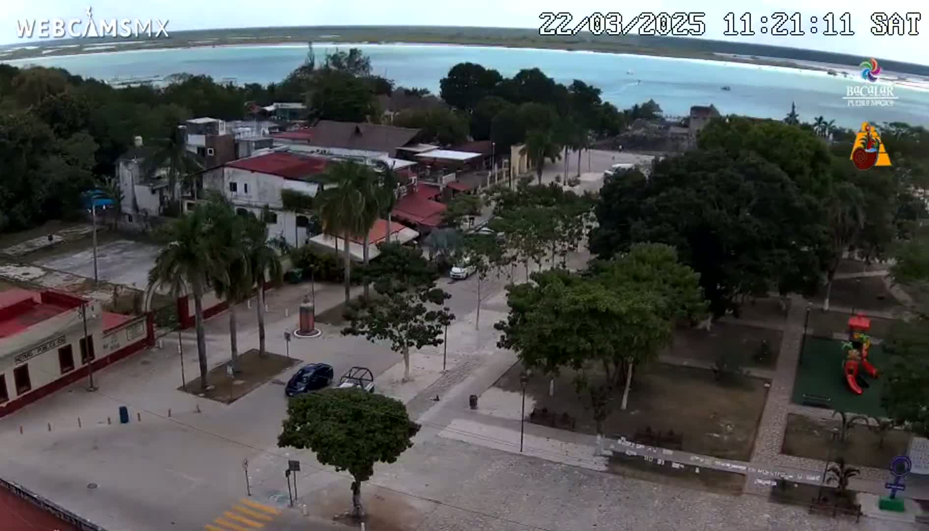 An aerial view of a town square with buildings, trees, and a playground, overlooking a calm body of water under an overcast sky.