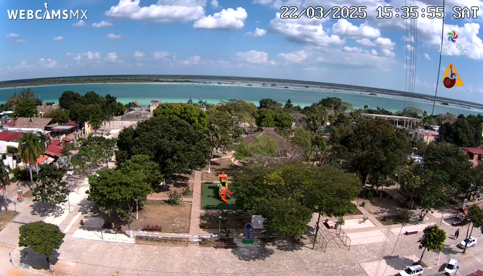 An elevated view of a town square with a playground, surrounded by lush green trees and buildings, overlooking a calm, turquoise body of water under a partly cloudy sky.