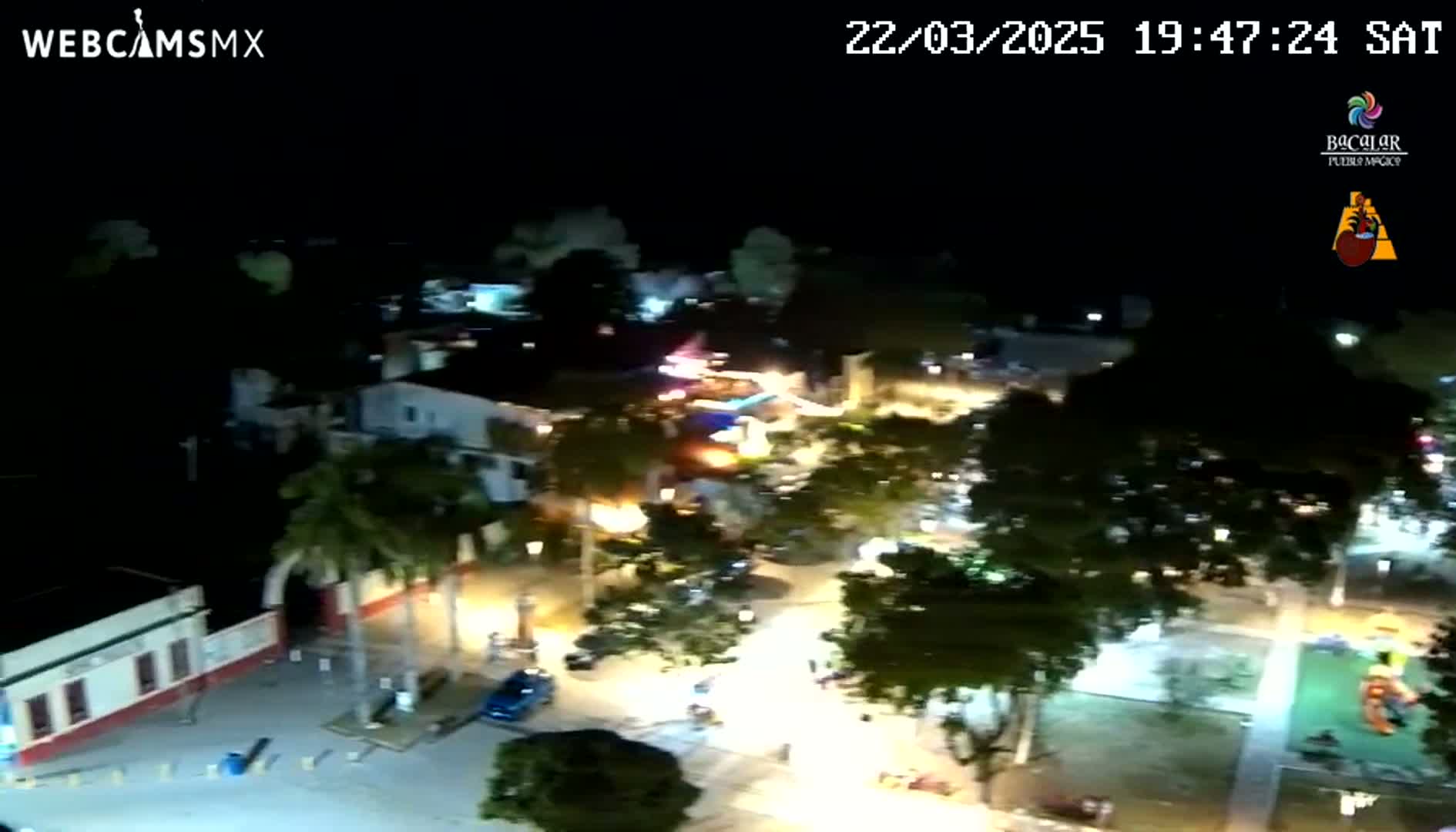 An aerial nighttime view shows a town square with illuminated buildings, trees, and vehicles under a clear, dark sky.