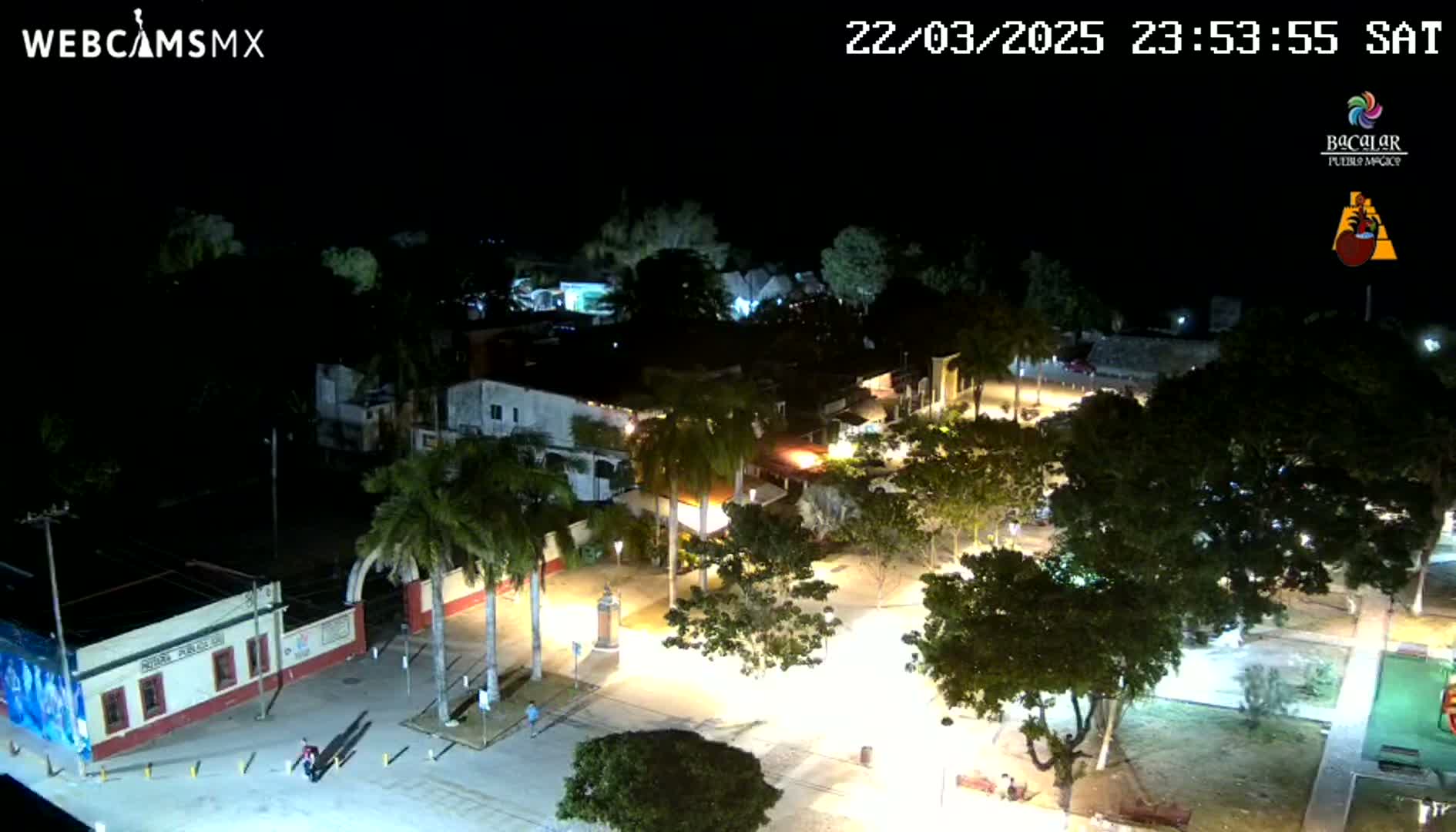 A nighttime aerial view of a town square, illuminated by streetlights, with palm trees and buildings surrounding a plaza.