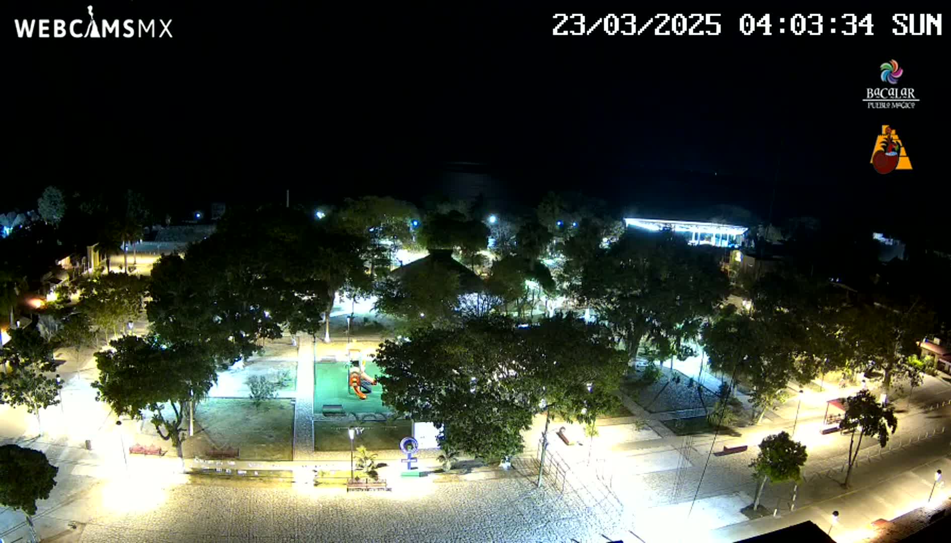 An illuminated park and plaza at night, featuring trees, pathways, and playground equipment.