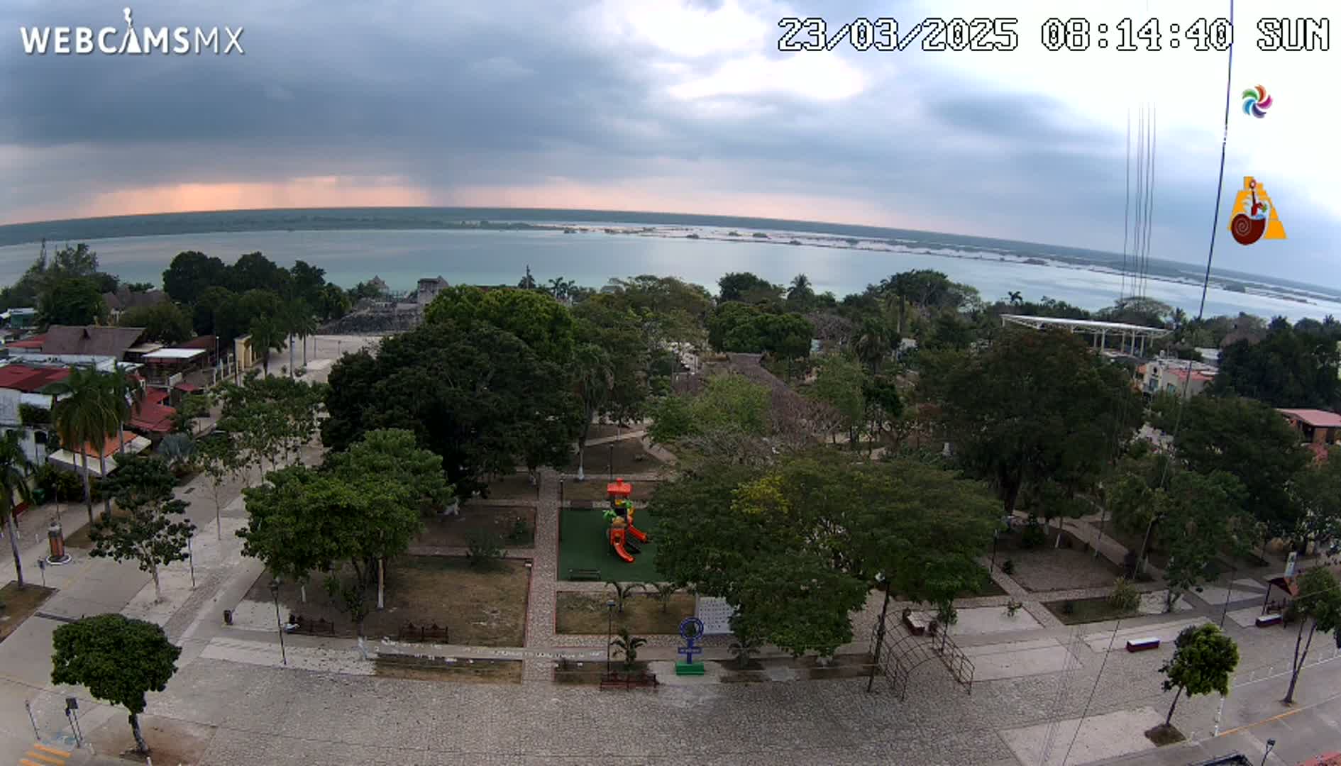 An overcast day reveals a town square with a playground, surrounded by lush green trees and buildings, overlooking a calm body of water.