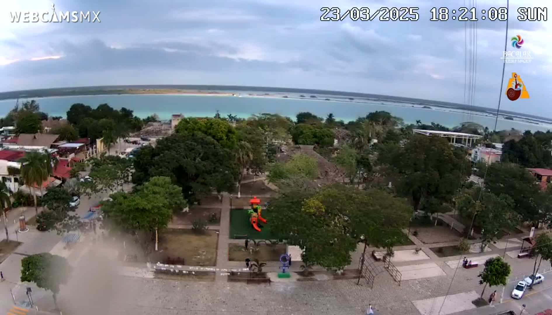 An aerial view of a town with a park and playground near a calm, turquoise body of water under a partly cloudy sky.