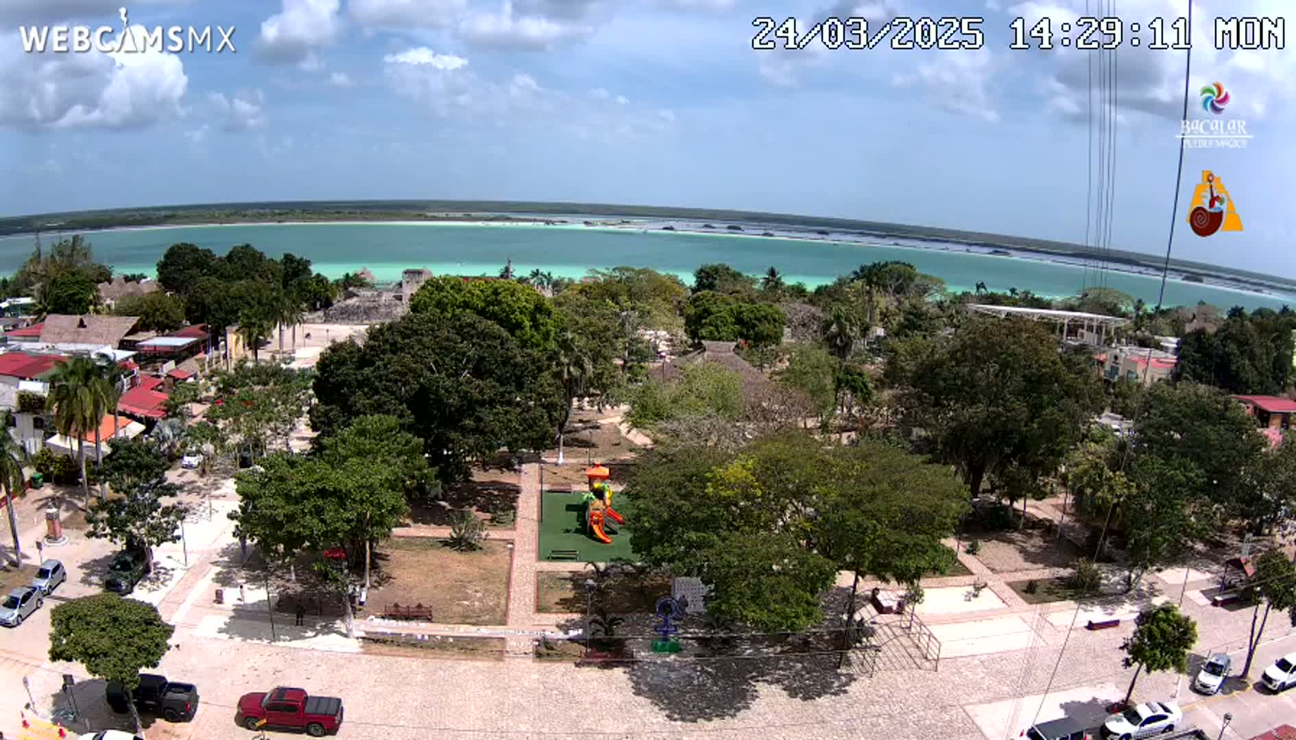 A park with a playground is situated near a town overlooking a calm, turquoise body of water under a partly cloudy sky.