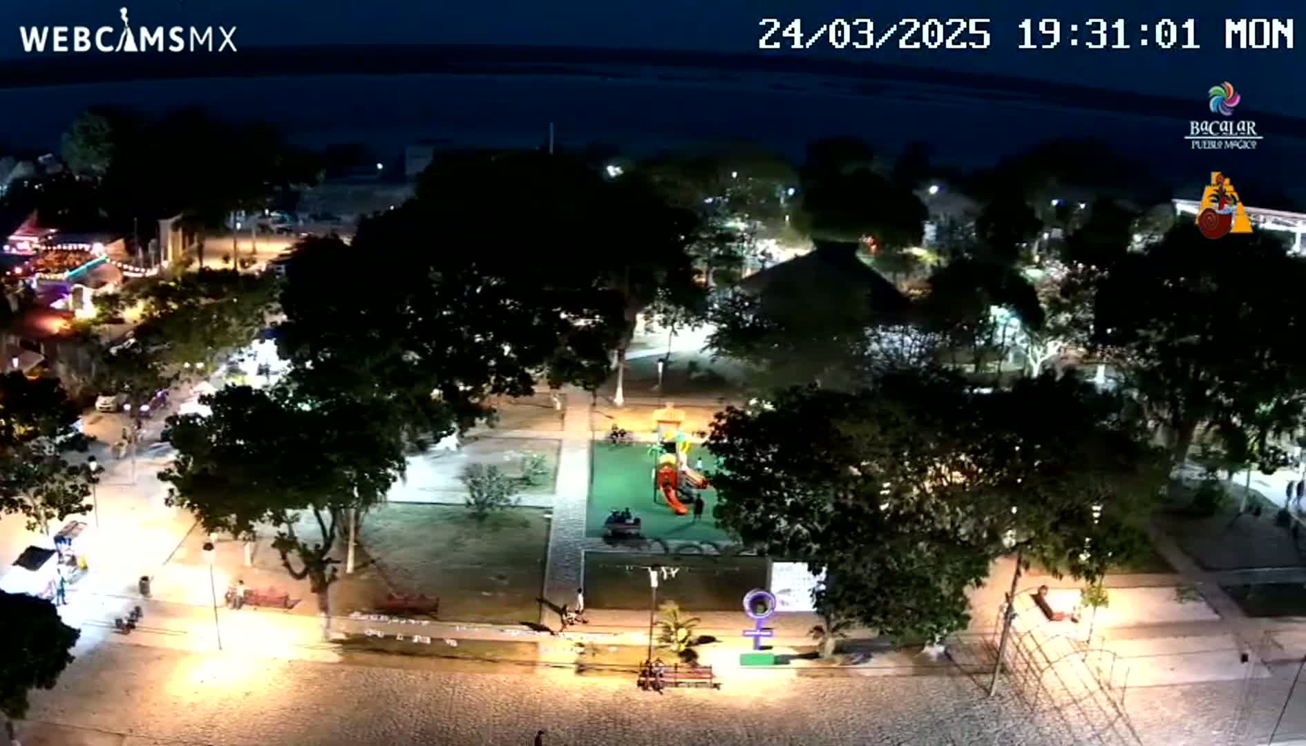 A nighttime aerial view shows a town square with a playground, trees, and walkways illuminated by lights.