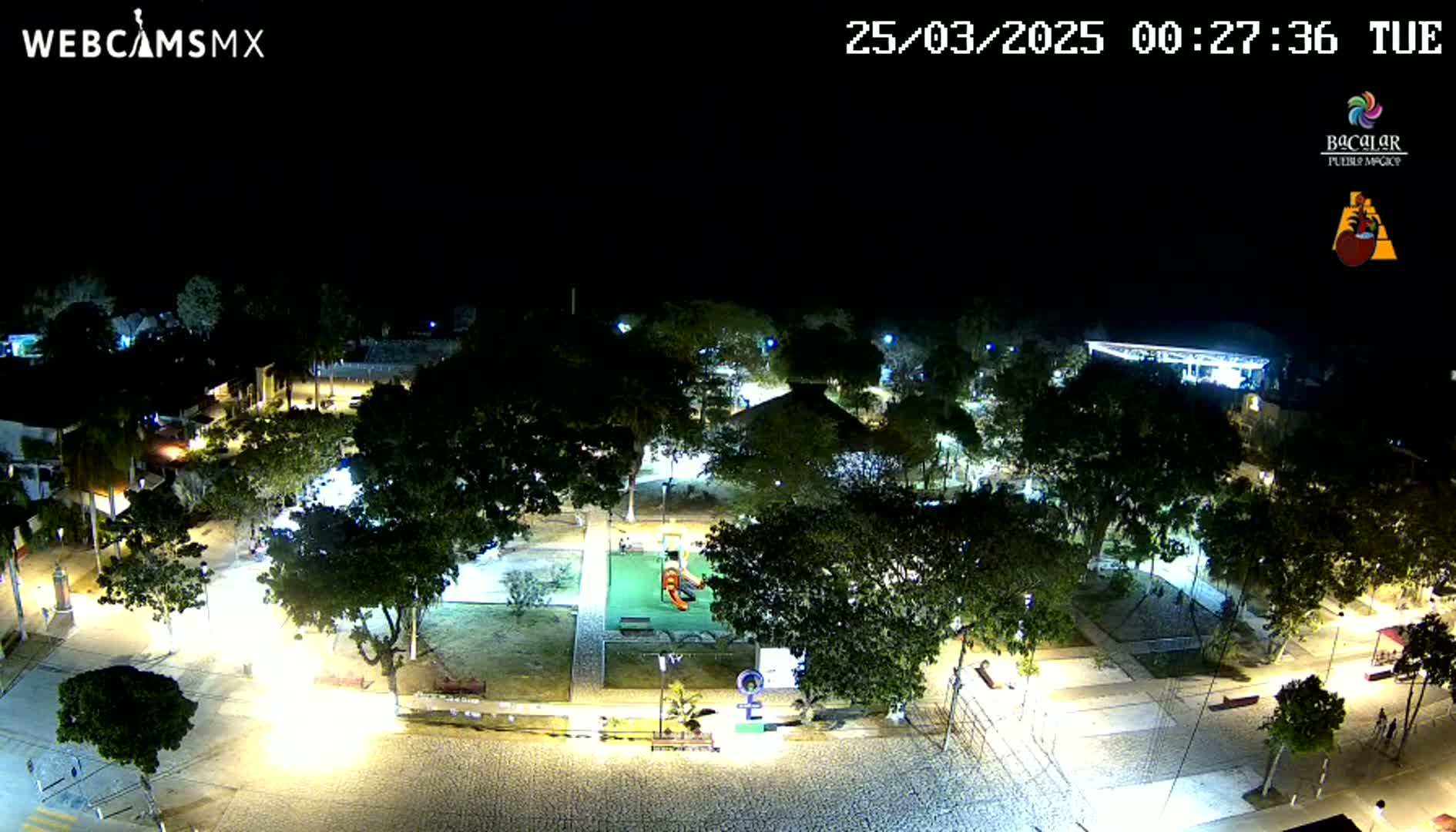 A nighttime aerial view shows a park with playground equipment, surrounded by trees and buildings, illuminated by streetlights under a clear, dark sky.