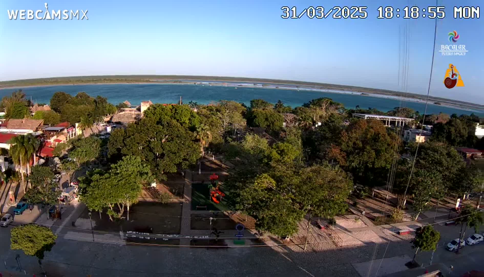 An aerial view of a town square with lush green trees and a playground, overlooking a calm, turquoise body of water under a clear, sunny sky.