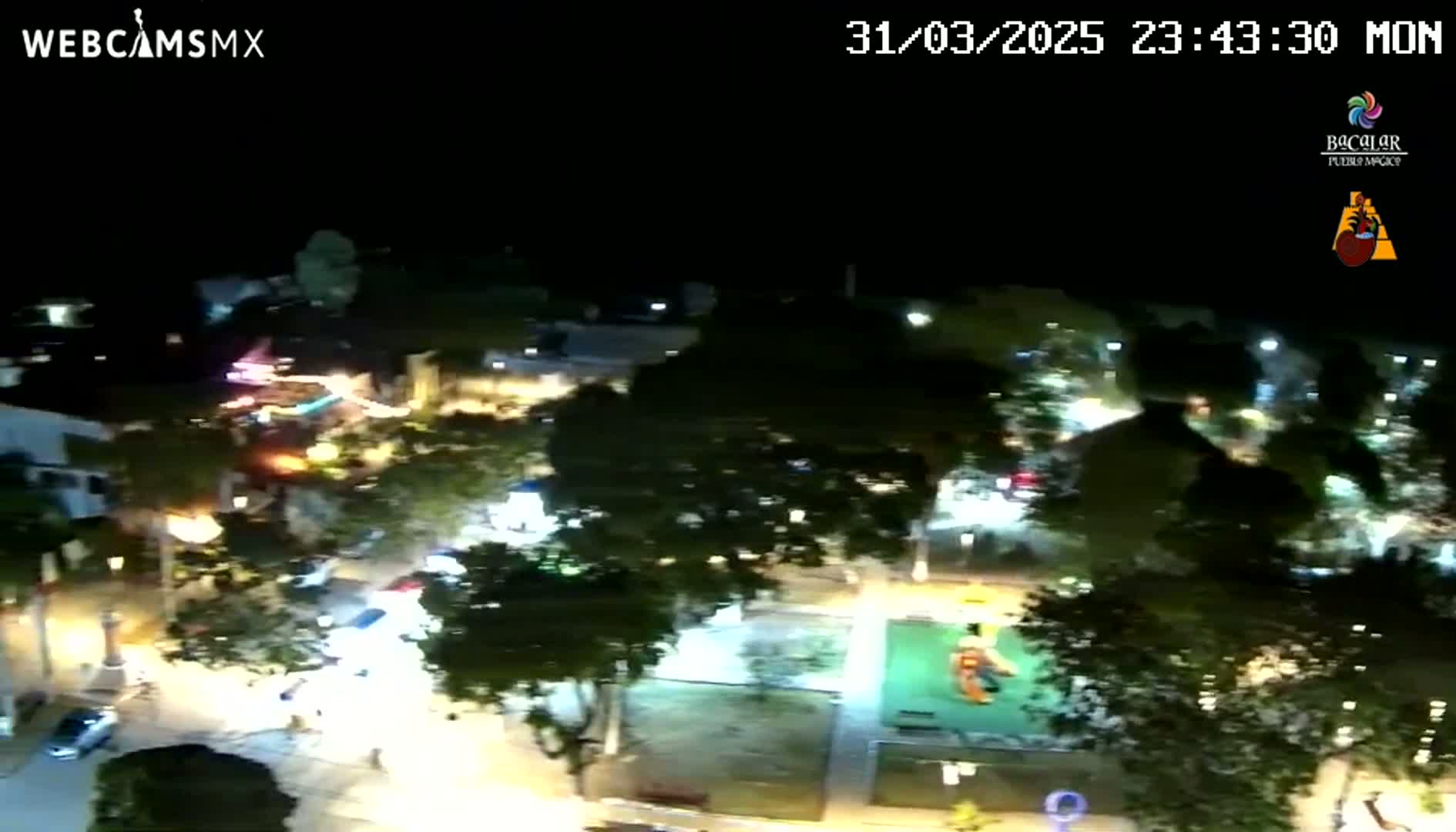 A nighttime aerial view shows a town square with streetlights illuminating cars, trees, and a playground.