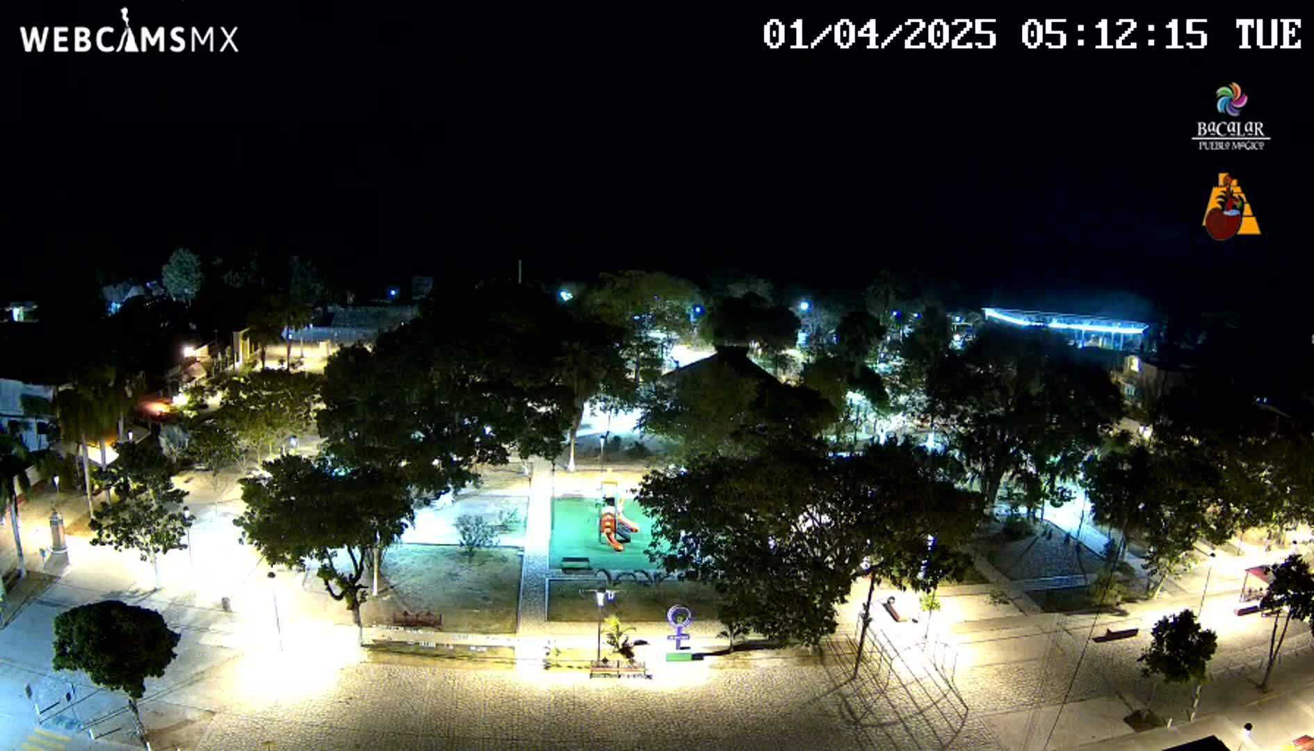 A nighttime overhead view of a park with illuminated pathways, trees, and playground equipment under a clear, dark sky.