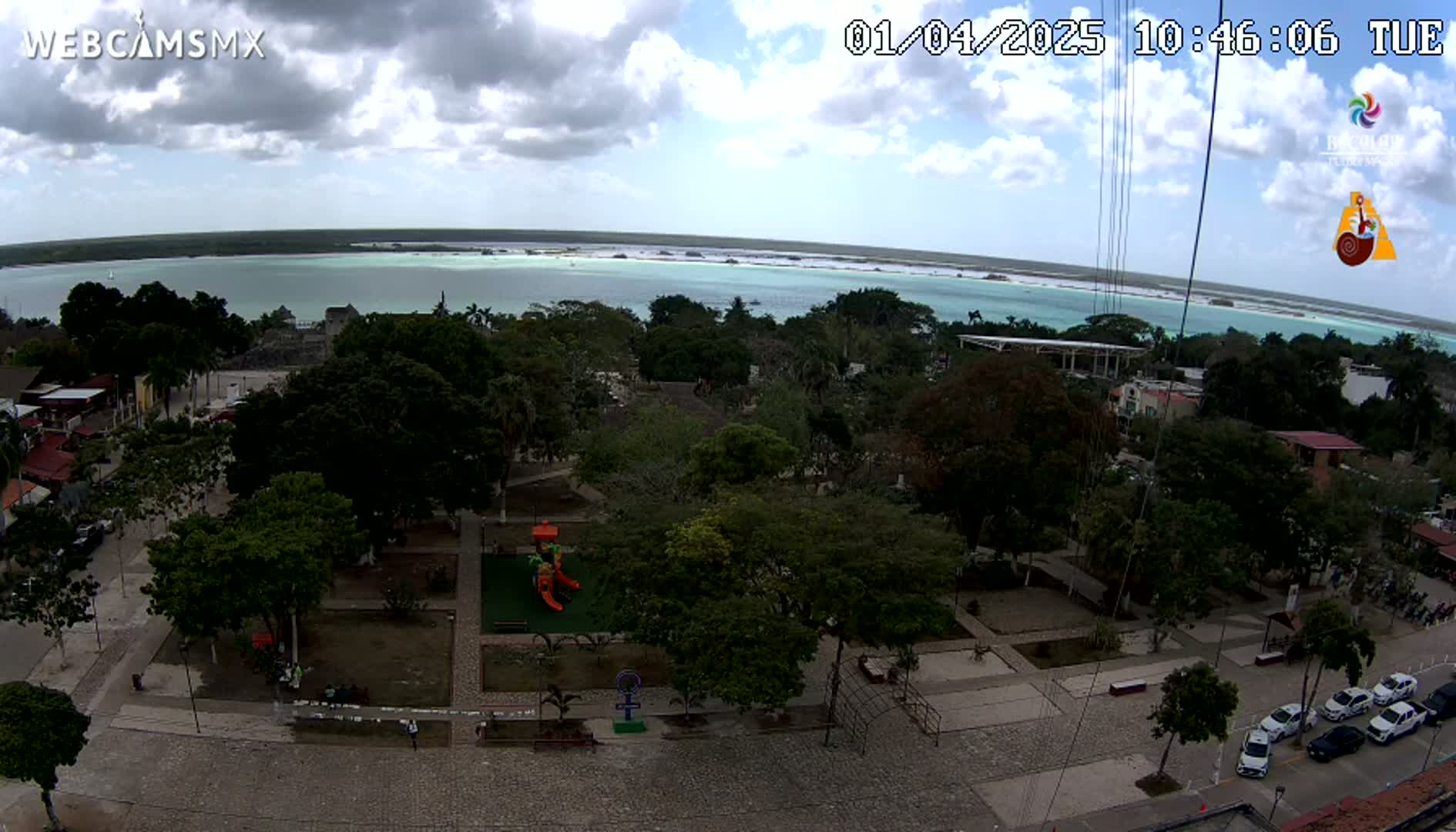 A partly cloudy day overlooks a town square with a playground and several parked cars, all situated near a body of calm, light-green water.