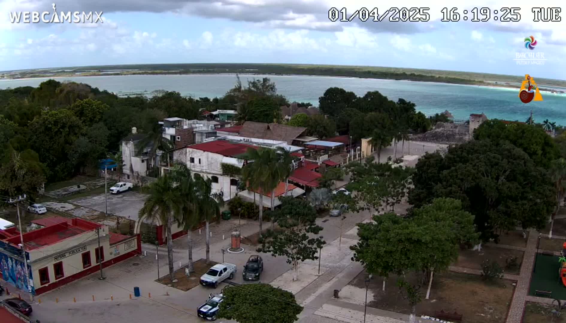 An aerial view of a town square with parked cars, palm trees, and buildings, overlooking a calm body of water under partly cloudy skies.