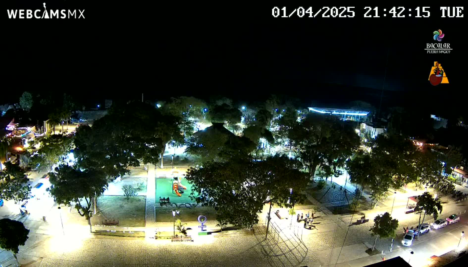 A nighttime aerial view of a town square with numerous trees, illuminated walkways, and a playground, under a clear, dark sky.