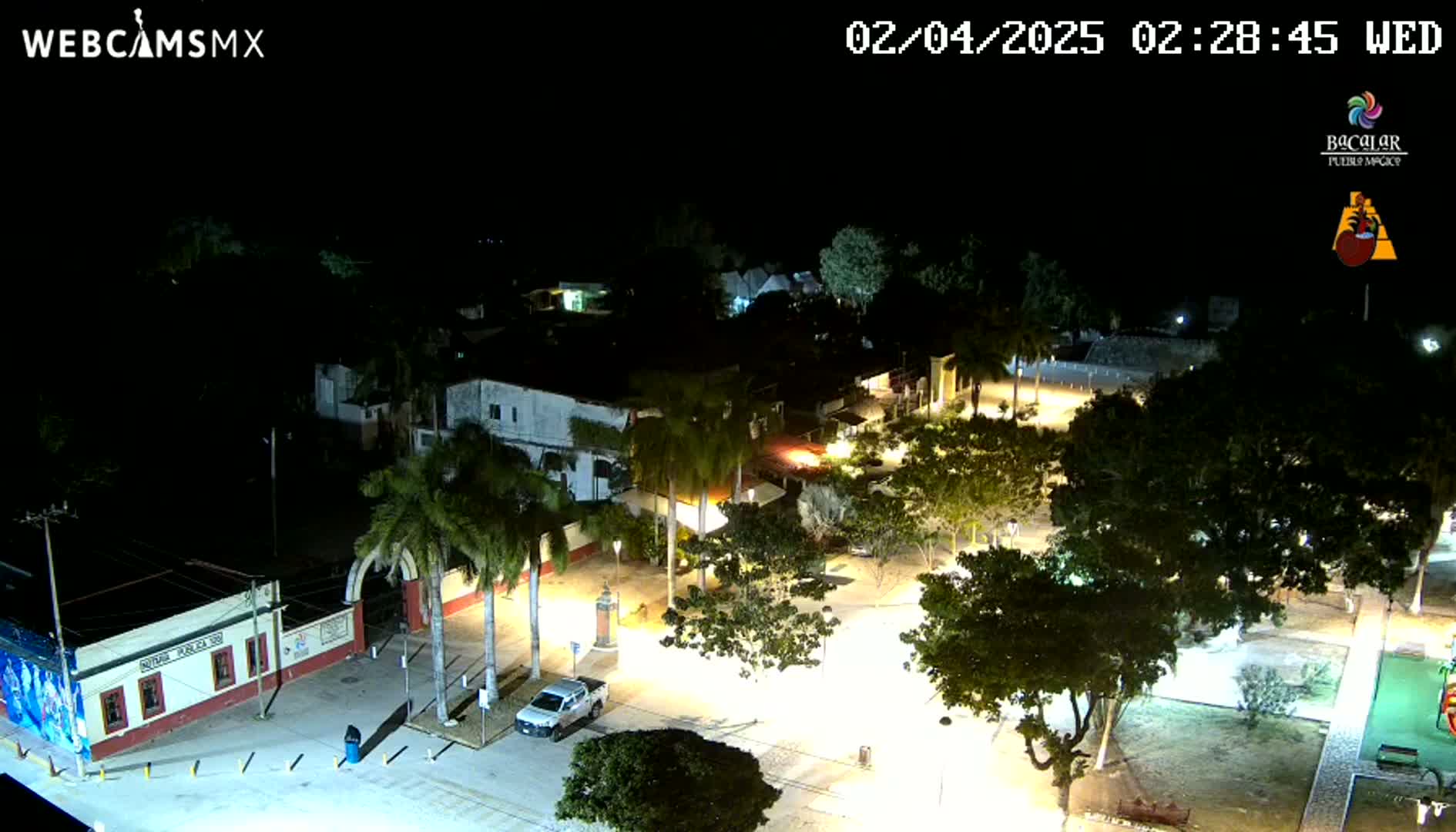 A nighttime aerial view of a town square with palm trees and buildings illuminated under clear skies.