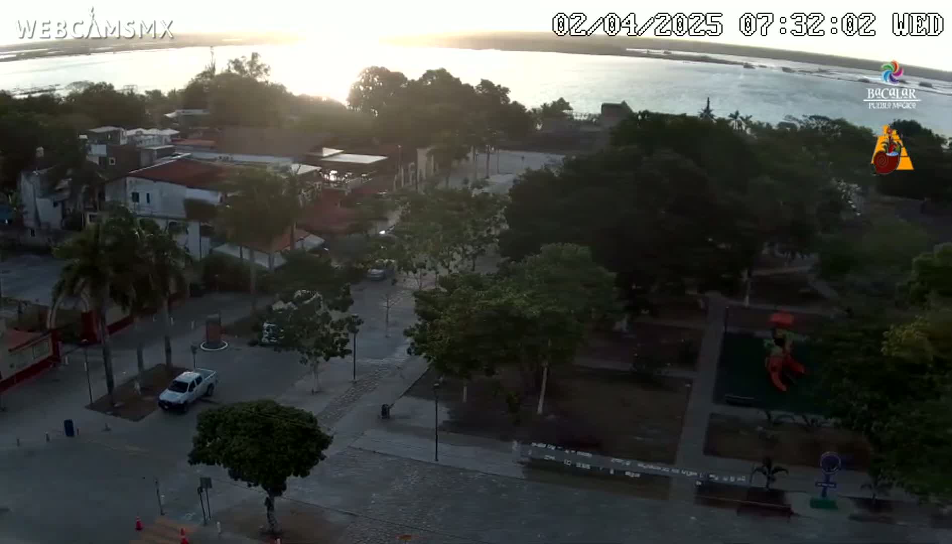 An aerial view shows a town square with parked cars, trees, and a playground, overlooking a calm body of water at sunrise.