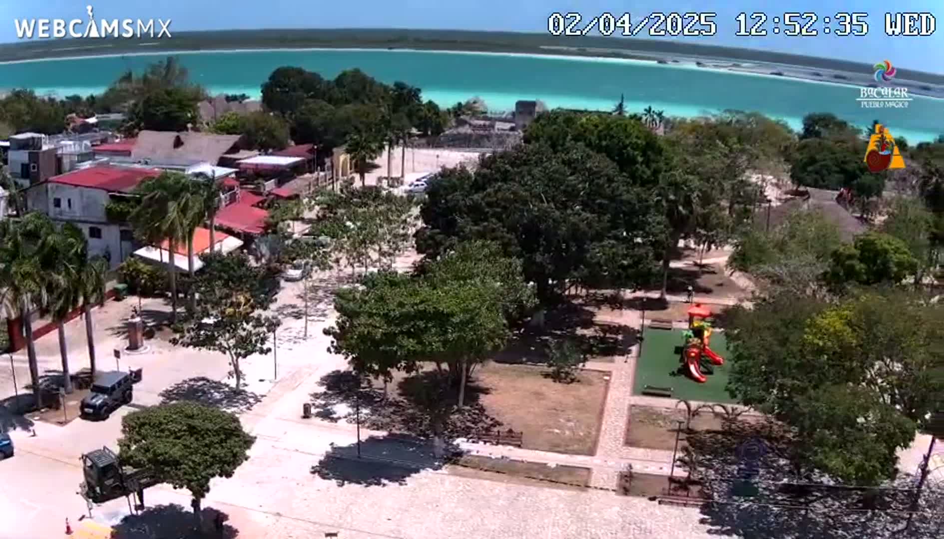 An elevated view shows a town square with a playground and a turquoise body of water in the background on a sunny day.