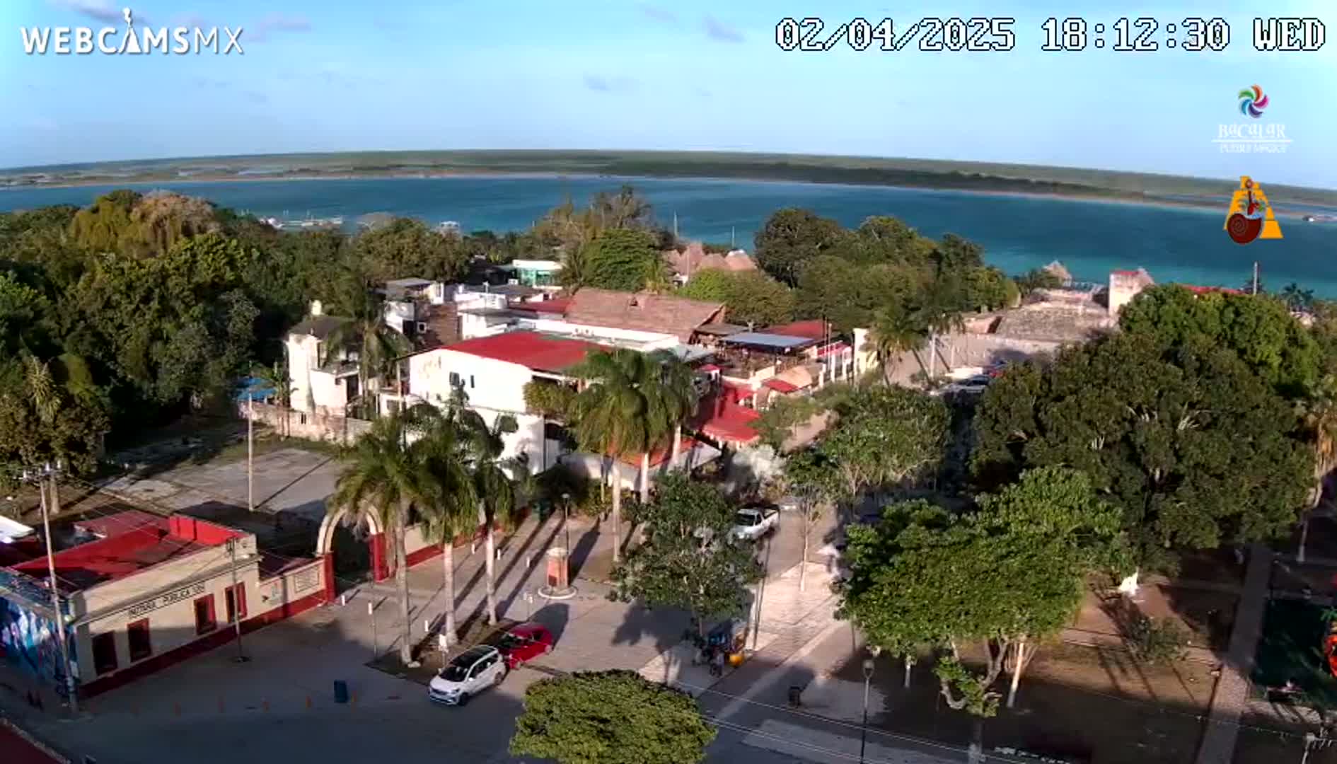 An elevated view of a town on a sunny day shows a clear turquoise body of water in the background,  palm trees and buildings in the foreground, with cars parked on the streets.