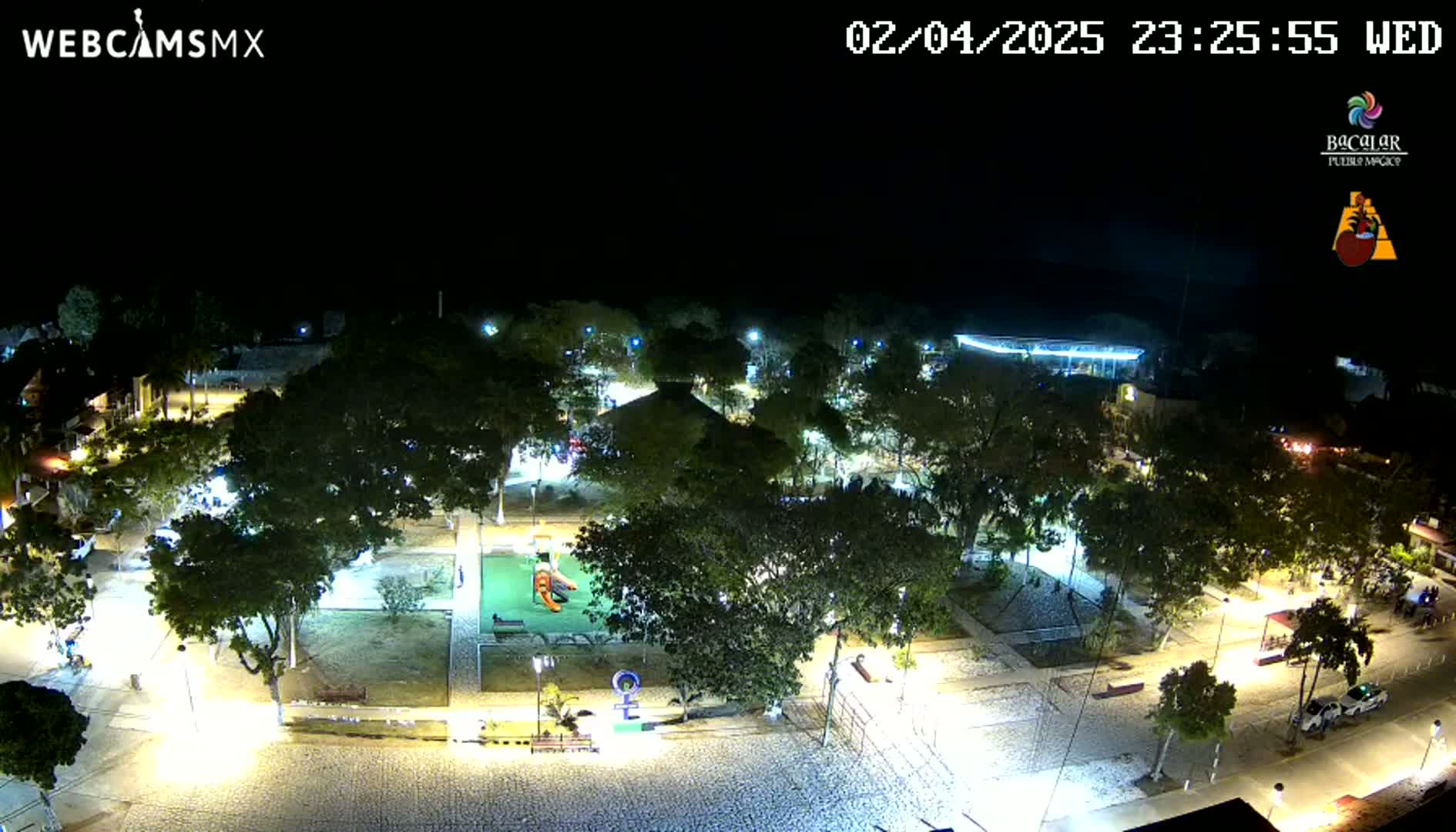 A nighttime aerial view of a town square with a playground, surrounded by trees and illuminated by streetlights under a clear, dark sky.