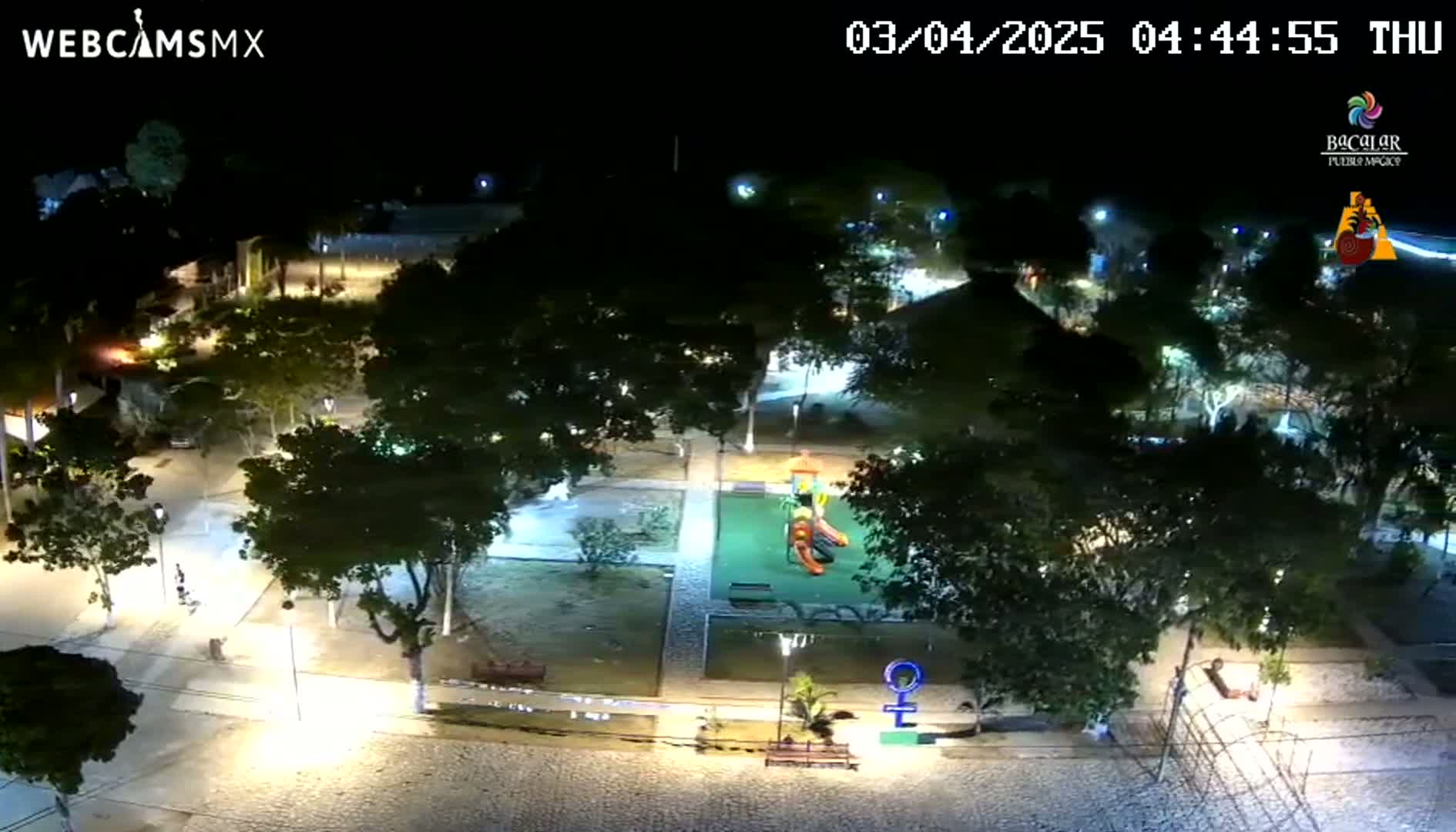 An illuminated park with a playground at night, surrounded by trees and paved pathways.