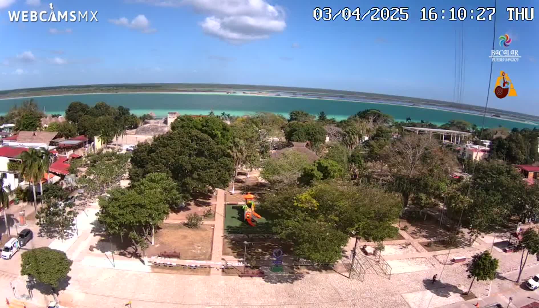 A town square with a playground is shown, next to a bright turquoise body of water under a sunny sky.