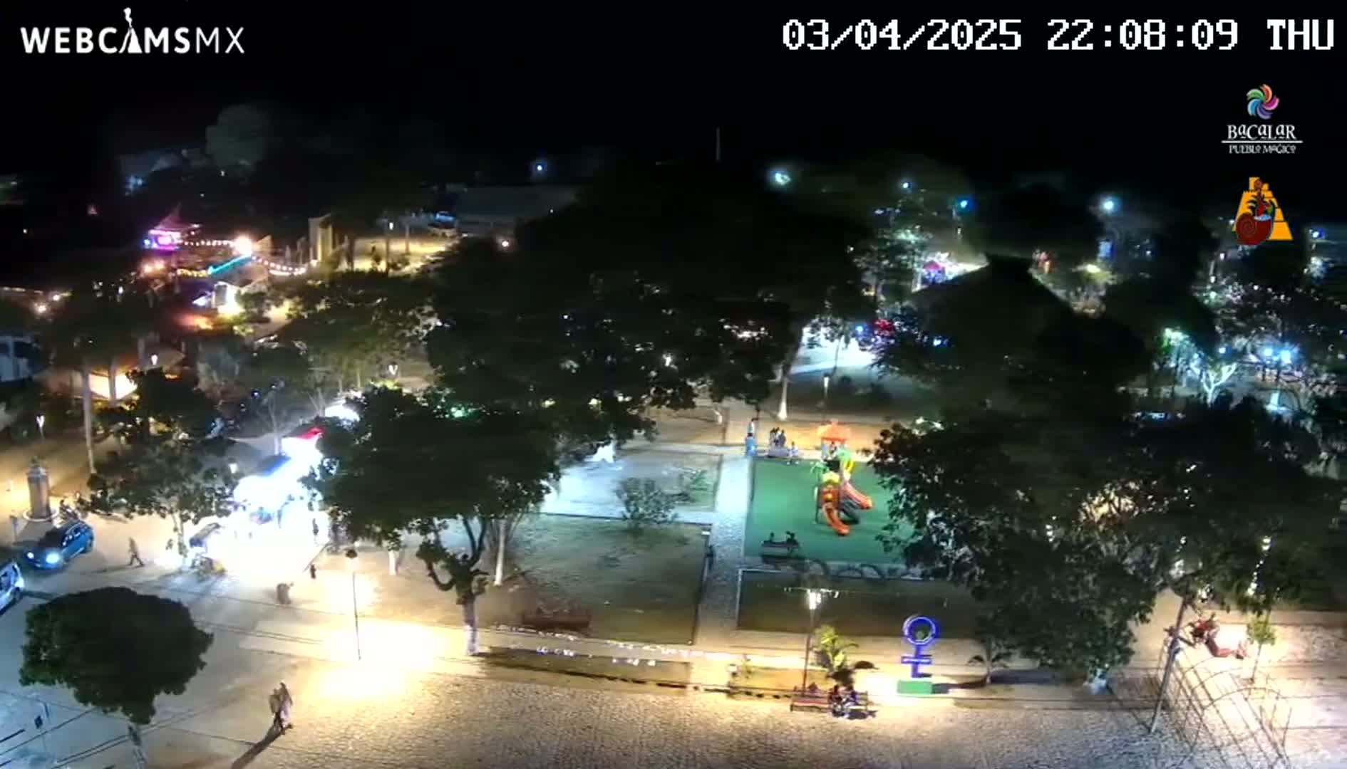 An aerial nighttime view of a town square with a playground, trees, and several people walking around, illuminated by streetlights.
